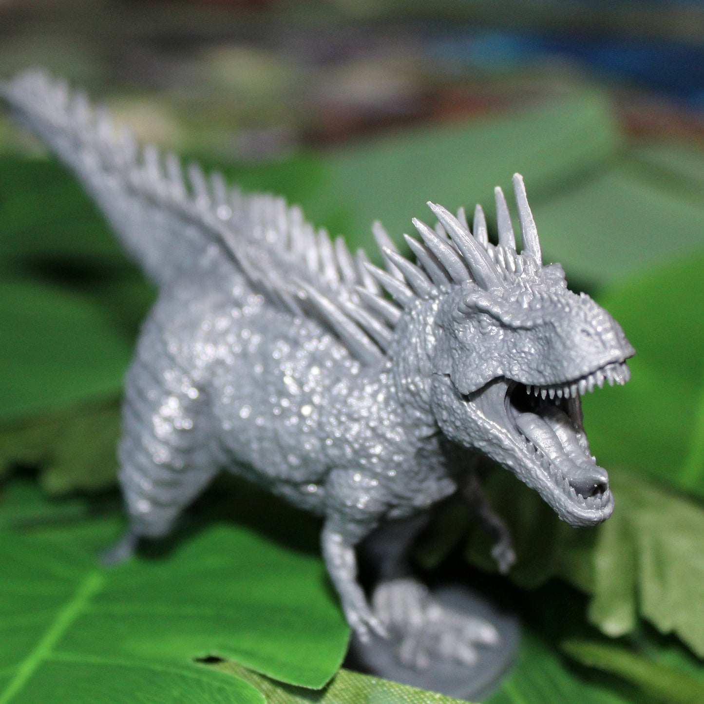 grey miniature figurine t-rex, mouth open and facing to right of the image. Standing on a pile of green leaves.