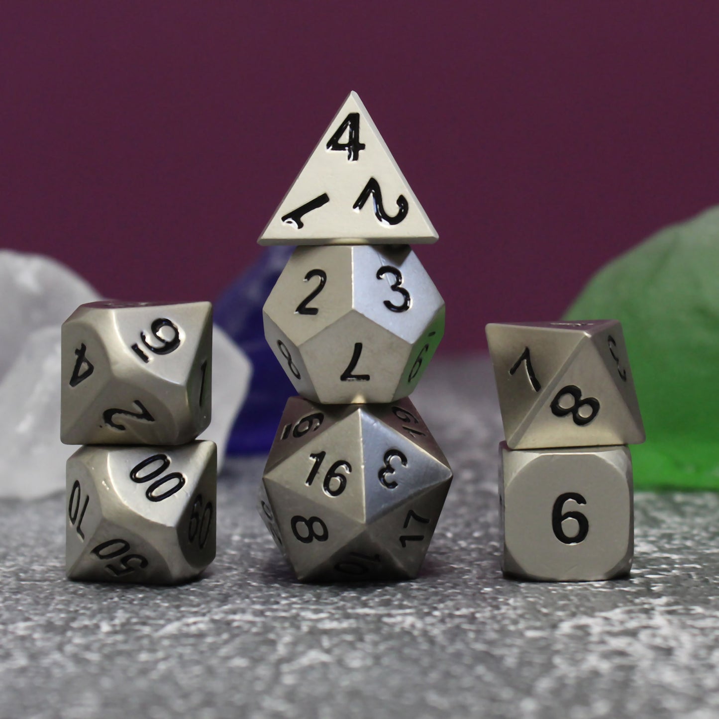 Bright silver dice set arranged in three stacks on a gray surface. Behind the dice are white, blue, and green stones, and a maroon background.