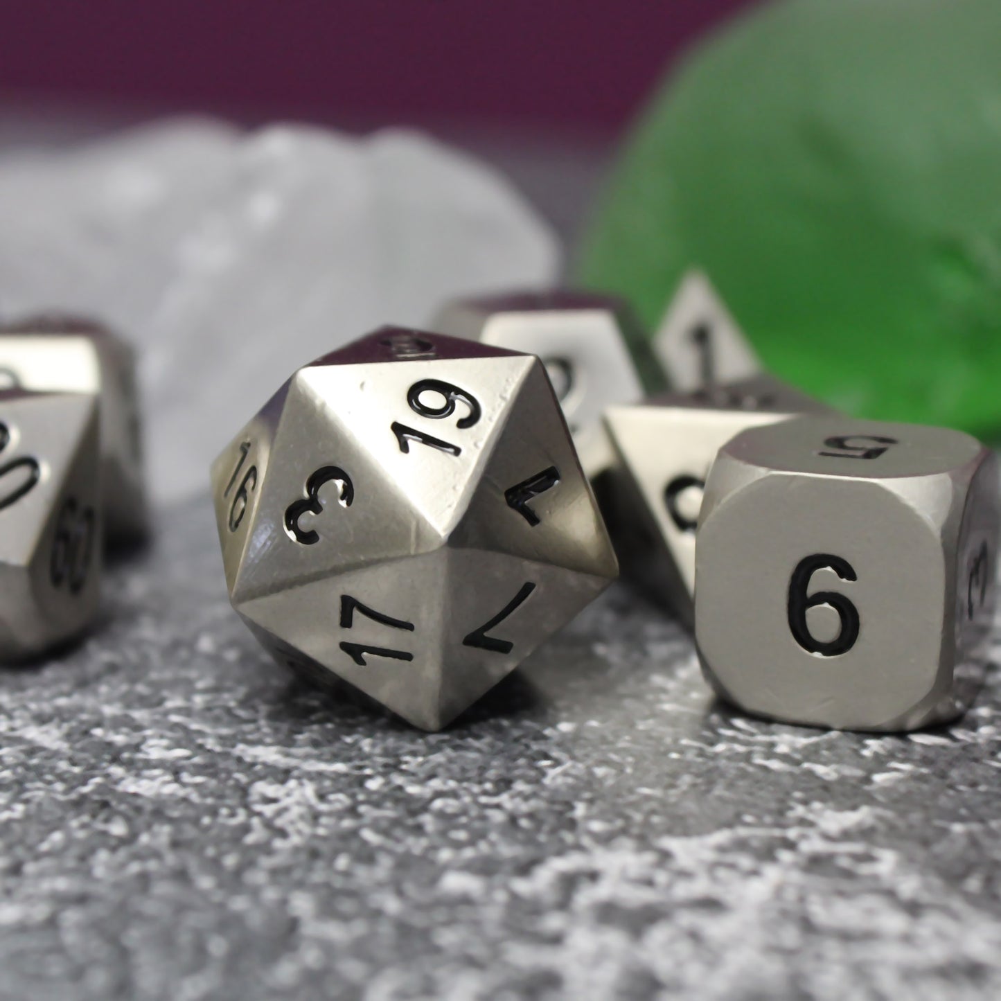 Close-up of the bright silver dice set on a gray surface. The close-up showcases the bright silver faces and black numbering. Behind the dice are white and green stones and a maroon background.