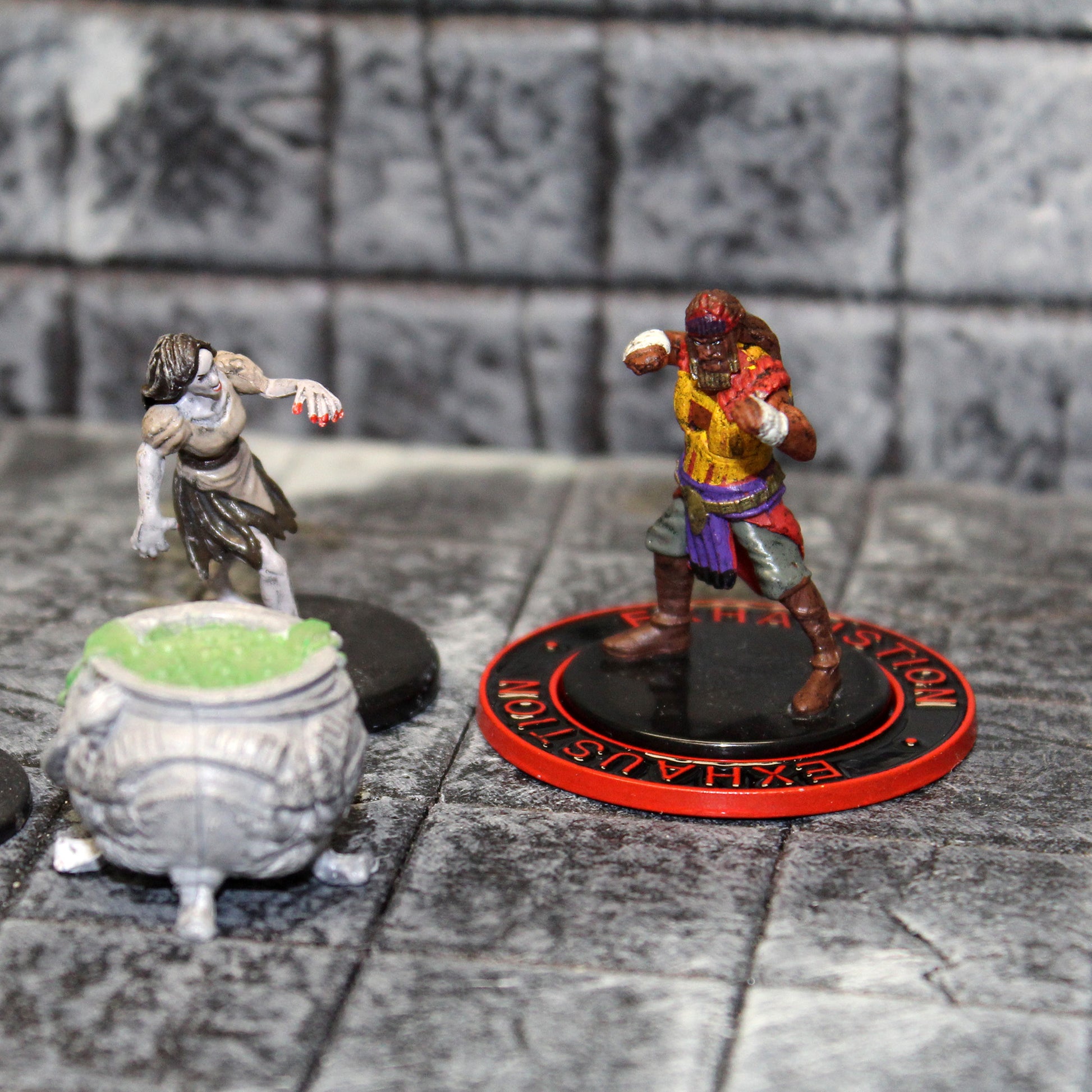 Two miniature figures, one with a black ring with a red rim and red text that says "Exhaustion" on it surrounding its base, sitting on a gray grid with a small cauldron sitting next to them.