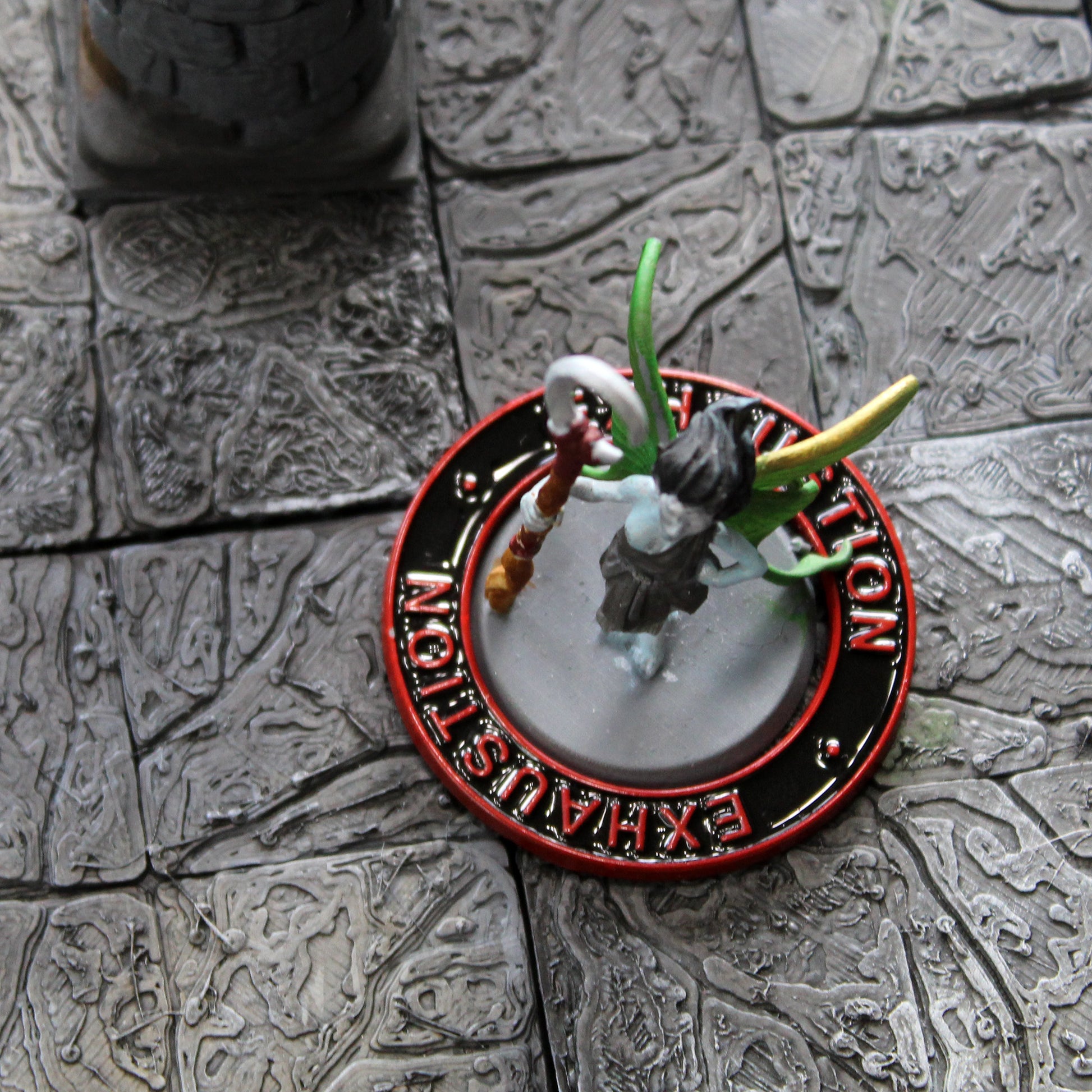 A figure of a winged fairy surrounded by a black ring with a red rim and red text that says "Exhaustion" on it on a gray grid.