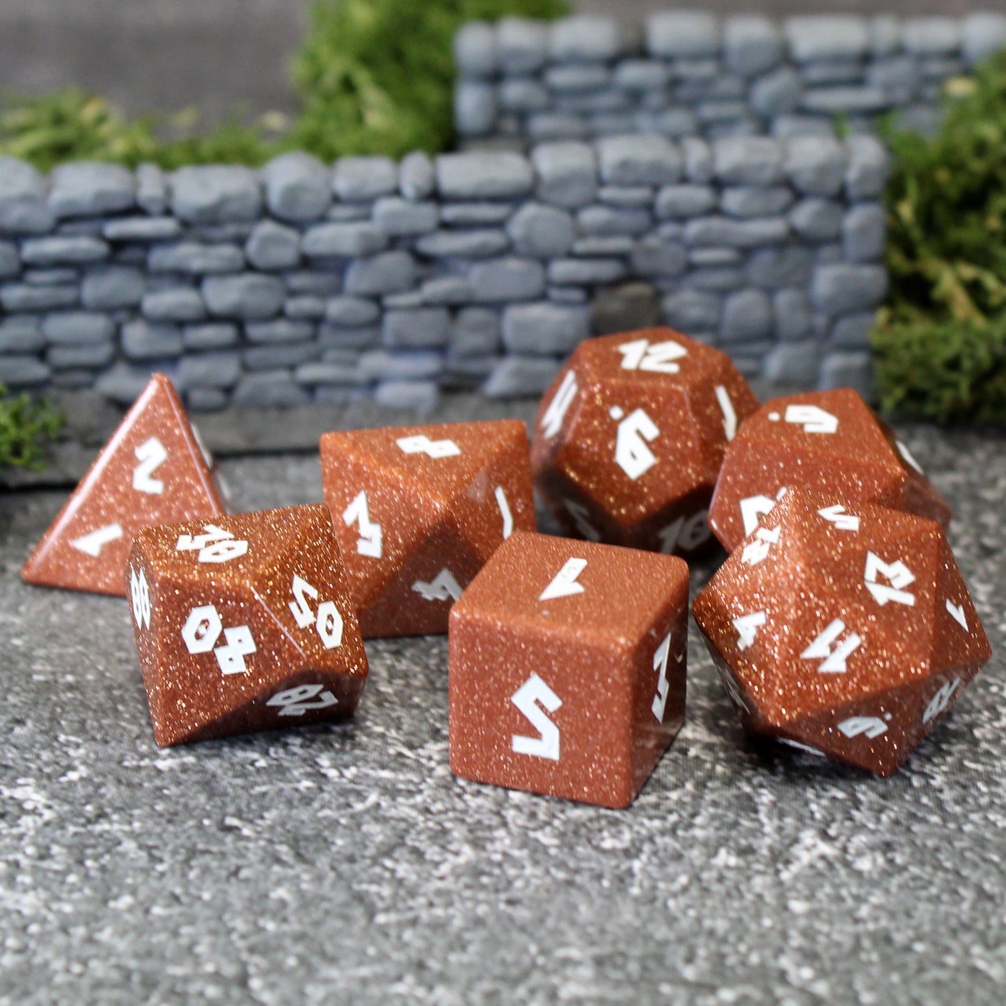 Goldstone dice set scattered on a pebble patterned play mat, with rock wall and foliage set pieces behind them