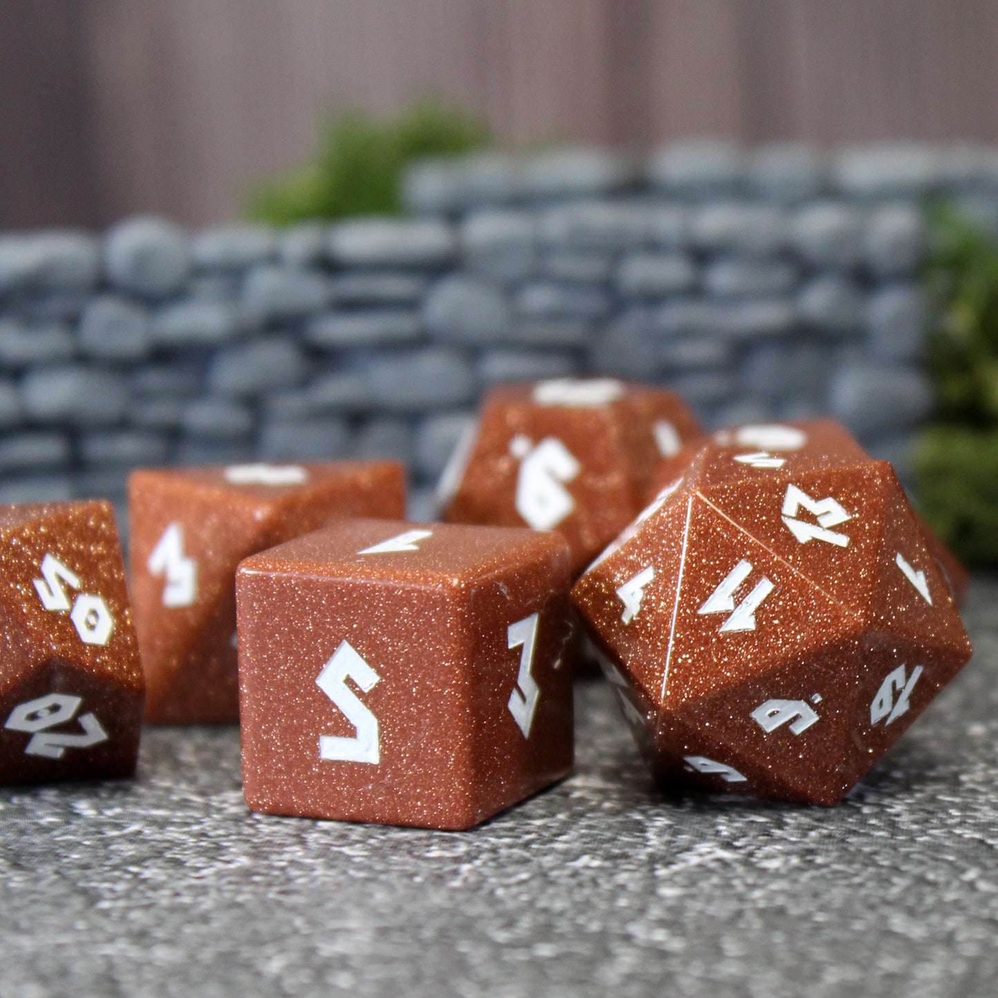 Goldstone dice set scattered on a pebble patterned play mat with a rock wall set piece behind them. The dice are a burnt orange color with glitter throughout, and have white numbering on each face.