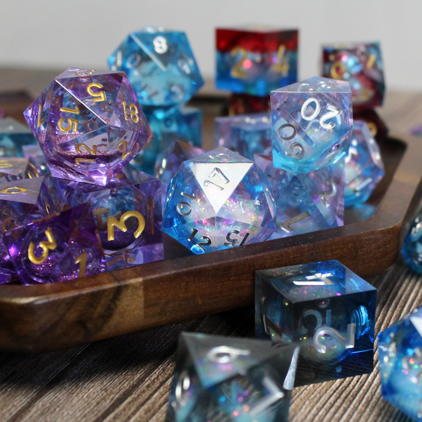 Close up of blue with white numbering and purple with gold number liquid core resin dice in a wooden tray. Each mylar bag contains a matching set.
