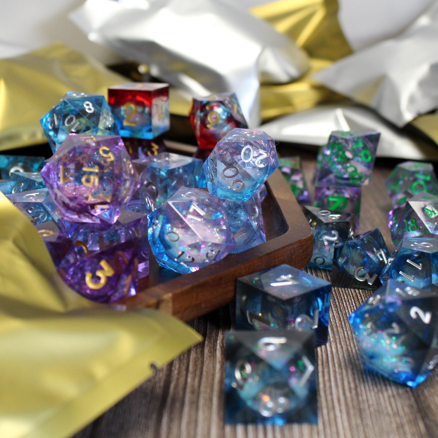 Display picture of liquid core resin dice arranged on a wooden surface with gold and silver mystery mylar bags in the background. Each mylar bag contains a matching 7-piece set of dice.