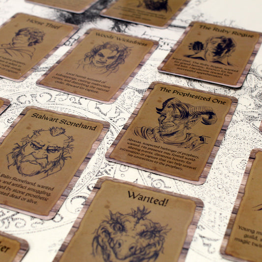 A arrangement of cards from the Lost & Wanted deck that showcases the format of each card having its own art.  Art is presented as a line art drawing with a description beneath it.
The central card has the text "The Prophesized One" above a picture of a smiling tiefling portrait, below the portrait reads, "Abraxas, suspected harbinger of prophecy, wanted.  His presence may foretell a world-altering events.  Generous bounty for information or capture that can help unravel his connection to the prophecy."