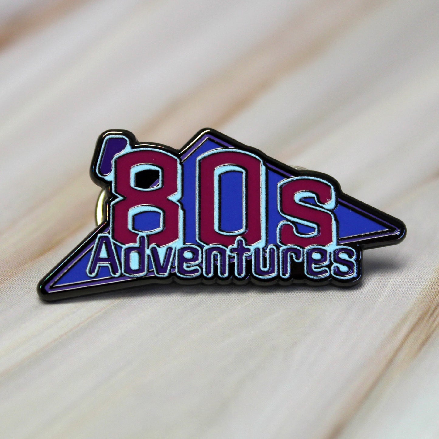 An "'80s Adventures" logo enamel pin sitting on a wooden surface.