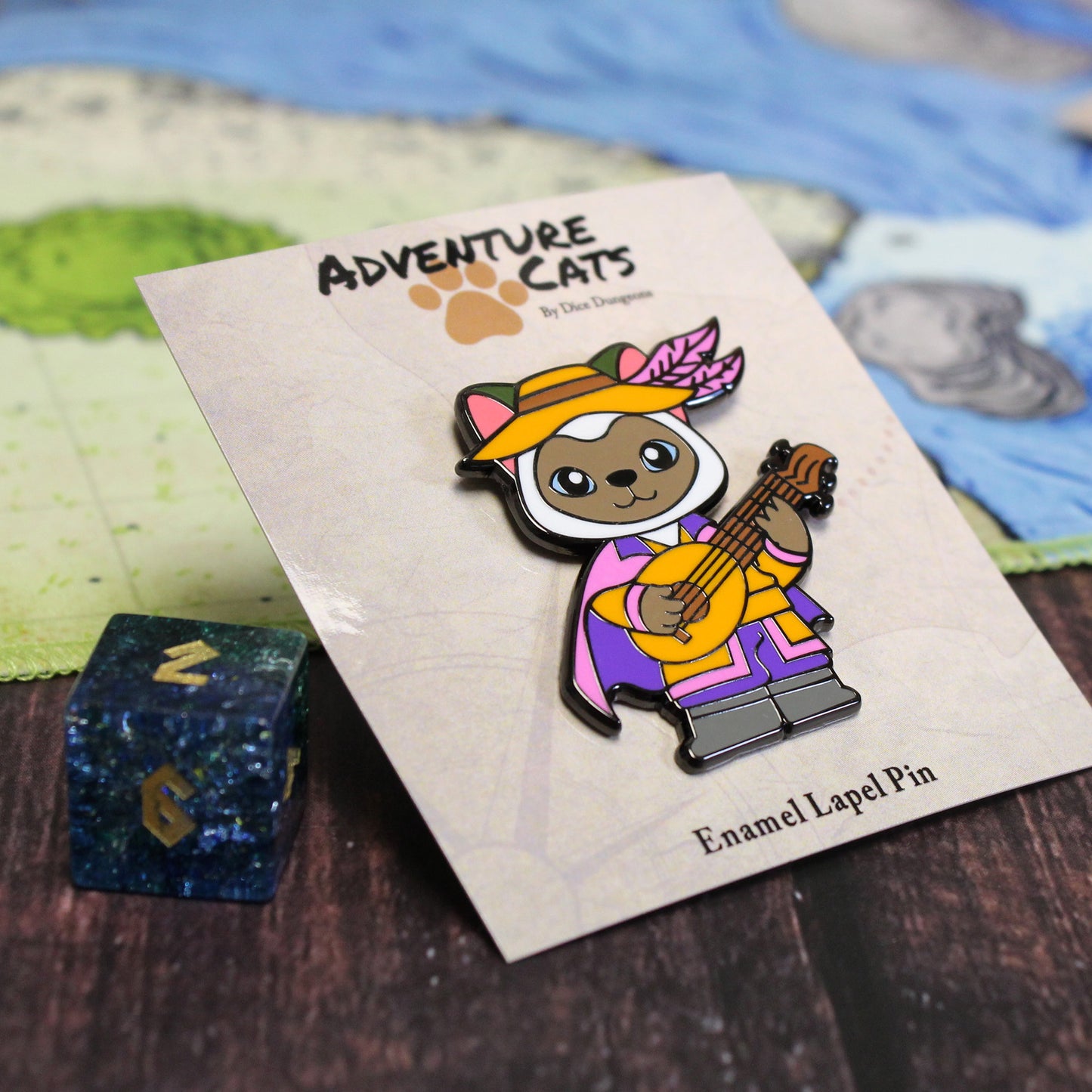 Adventure Cats bard enamel pin on a tabletop alongside its backing pin and a standard d6, providing a size comparison.