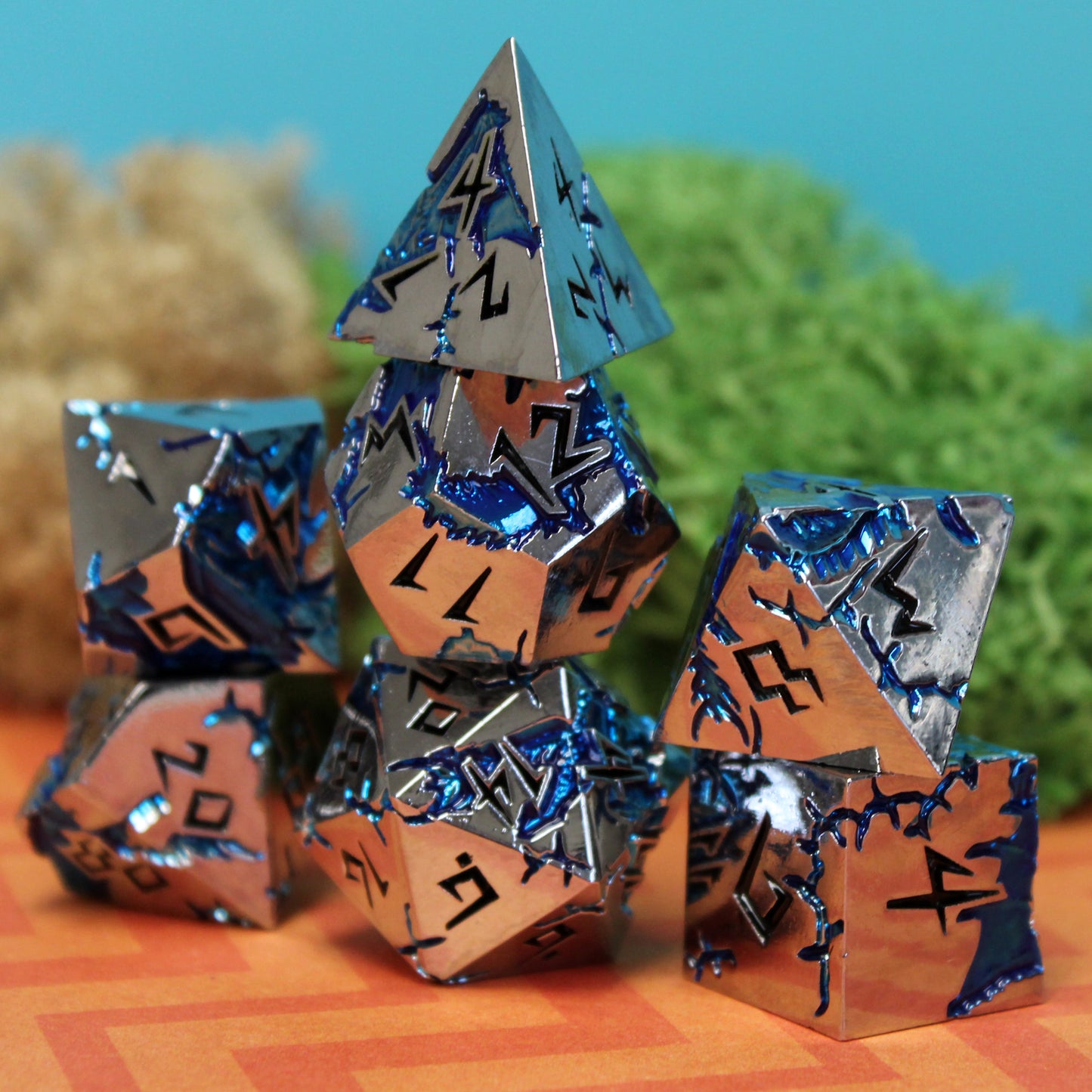 Stormrift silver metal dice set stacked in front of moss, featuring scarred blue enamel and black numbers, adding a natural backdrop for fantasy tabletop gaming.