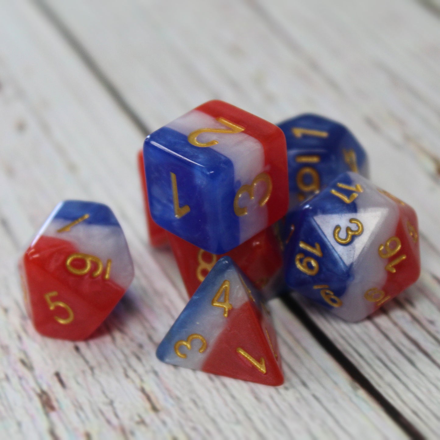 Full set of a red, white, and blue layered dice set on a wooden table, showing the gold numbering clearly on each d&d die face.