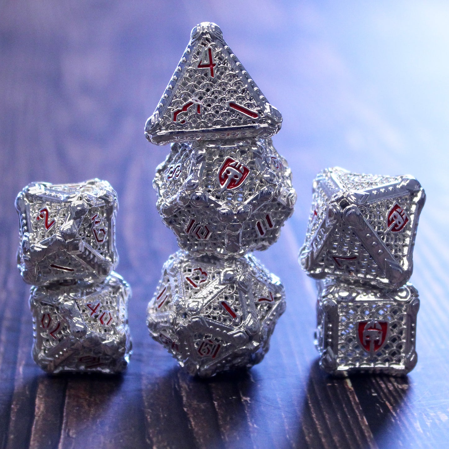 Stacked set of silver-colored fighter-inspired hollow metal RPG dice with a chain mail background on each die face, red numbers, and a helmet, sword, and shield replacing the high number on each die on a wooden surface