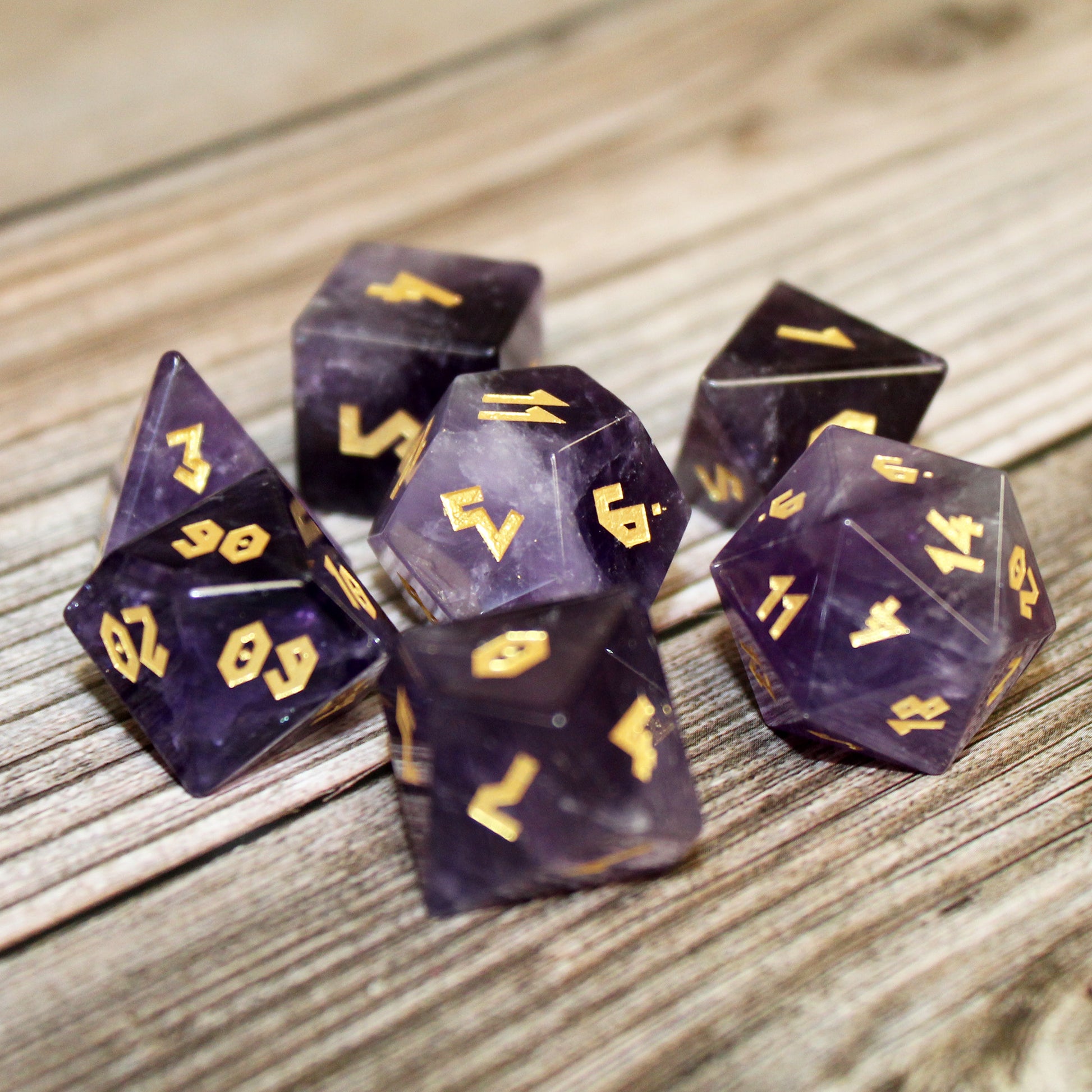 Amethyst gemstone dice set arranged on a wooden surface, with the gold Roman font numbering catching the light on the polished stone.