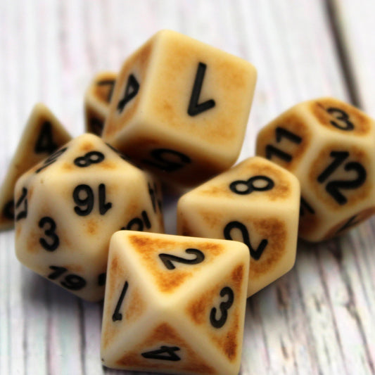 Close-up of scattered Ancient Bone Brown Polymer dice set on top of a white wooden surface. The dice have a bone appearance with black numbering.