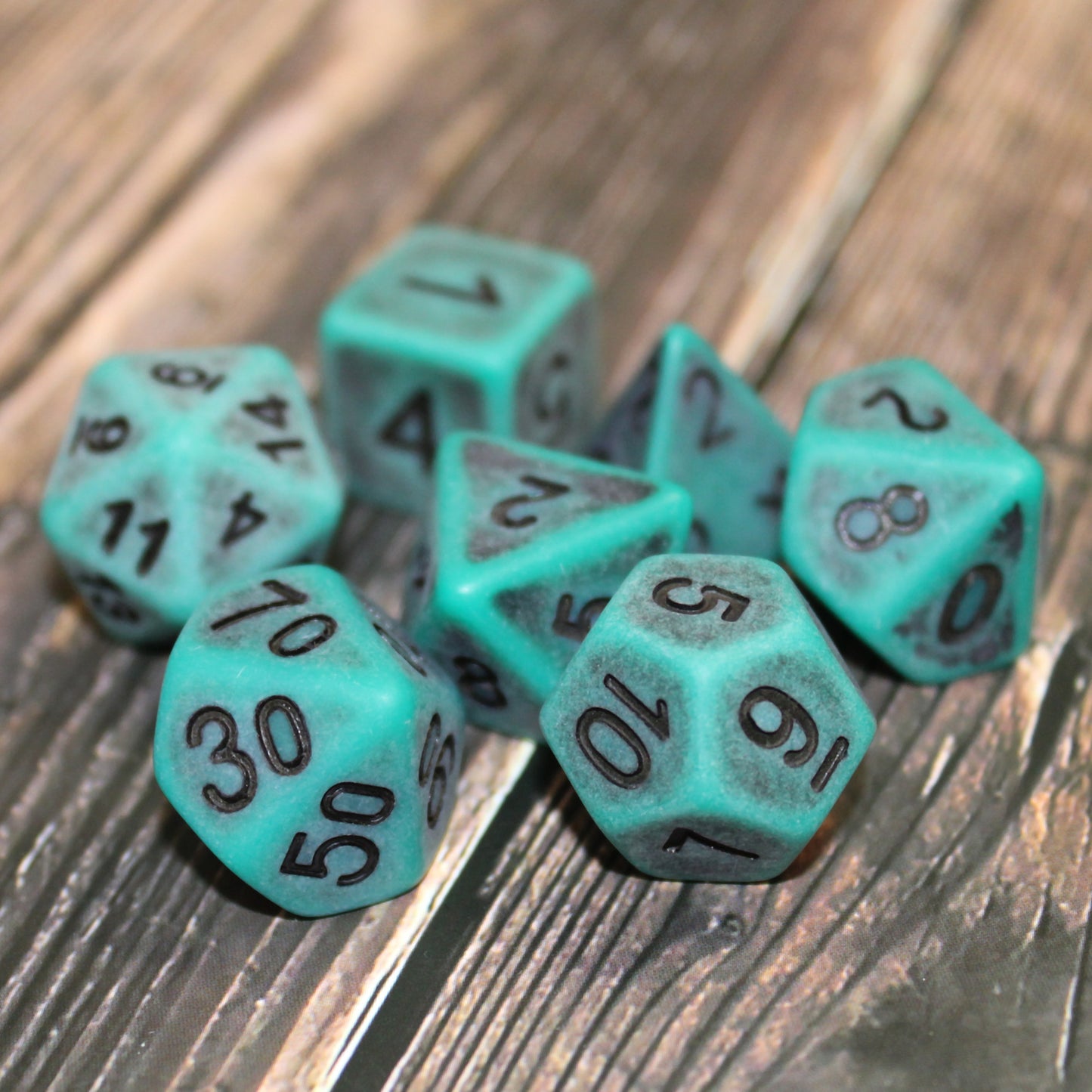 Closeup of a set of seven green plastic dice with black numbering designed to look aged on a wooden surface