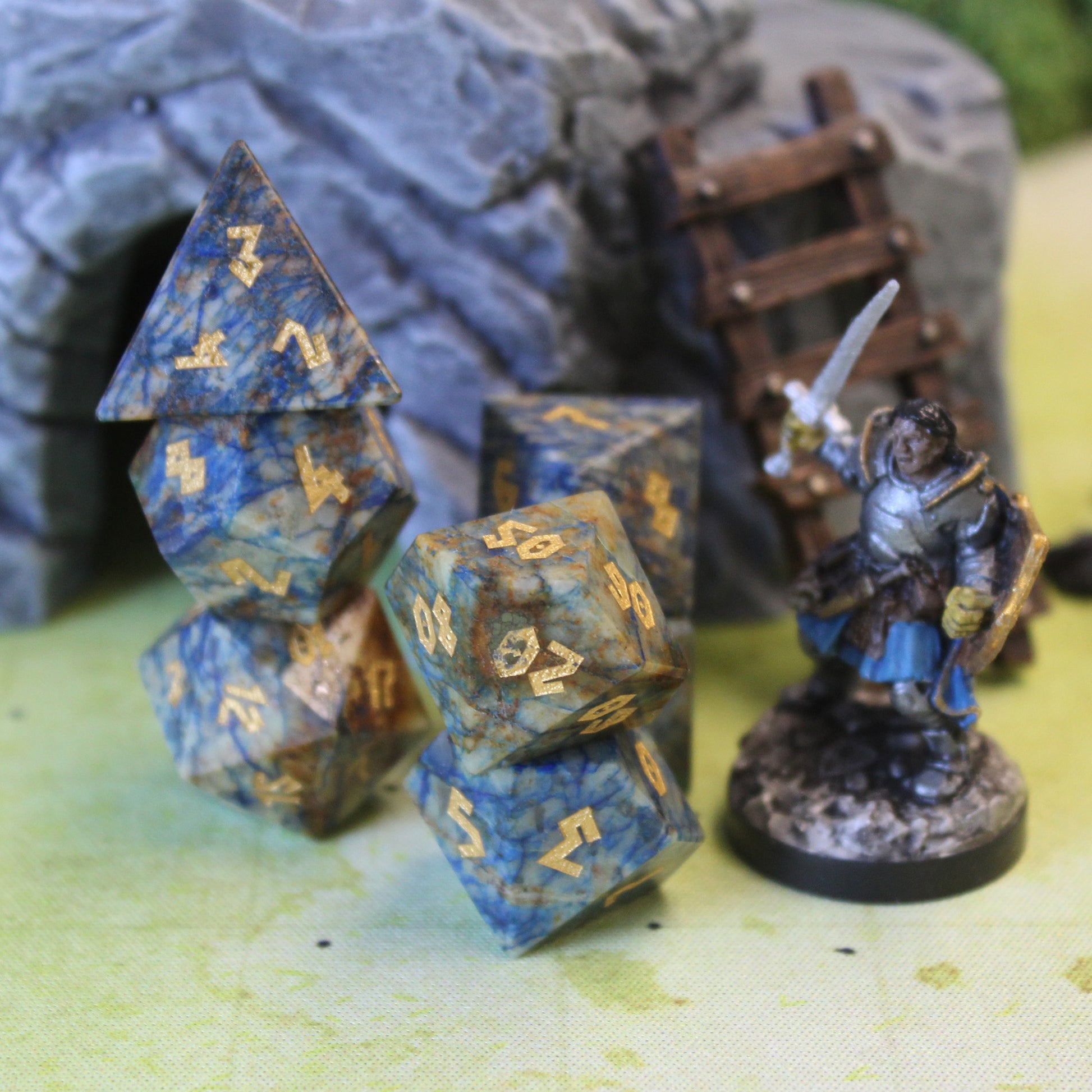 Azurite dice set displayed in three stacks on a green play mat. Next to the dice is a mini figure, and behind them is  a rocky cave opening set piece with a ladder leaning against it