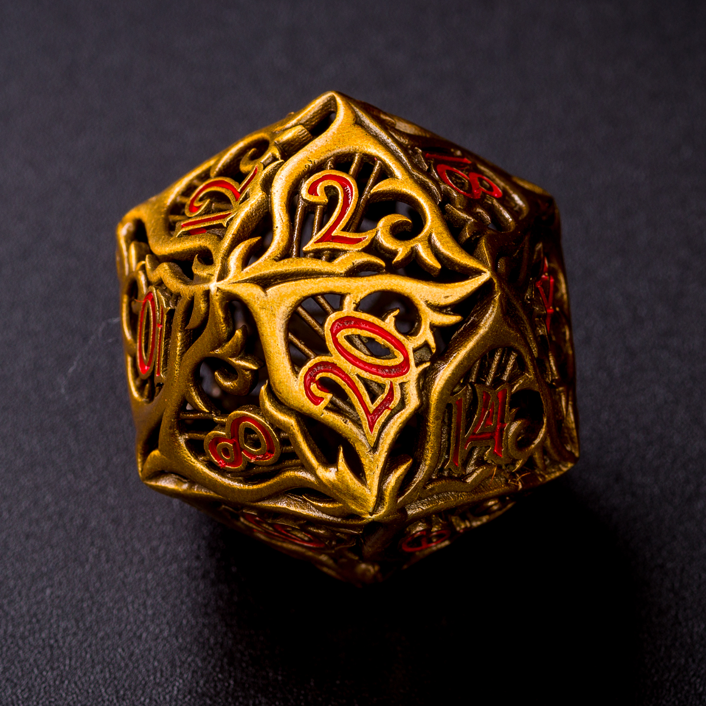 A closeup of a single gold-colored bard-inspired hollow metal twenty-sided die with red numbering and a hard motif on each face on a black background