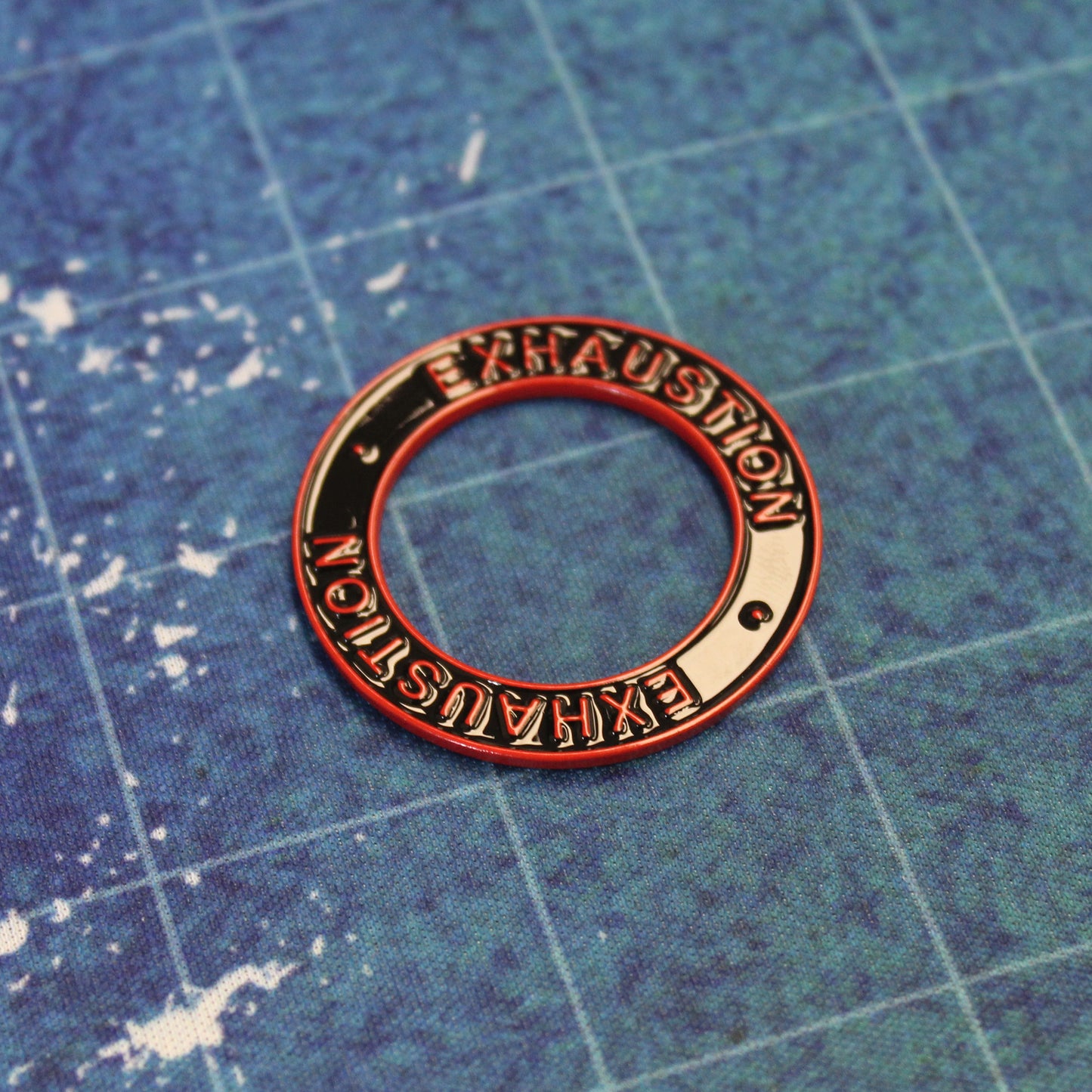 A black ring with a red rim and red text that says "Exhaustion" on it sitting on a blue gridded gaming mat.