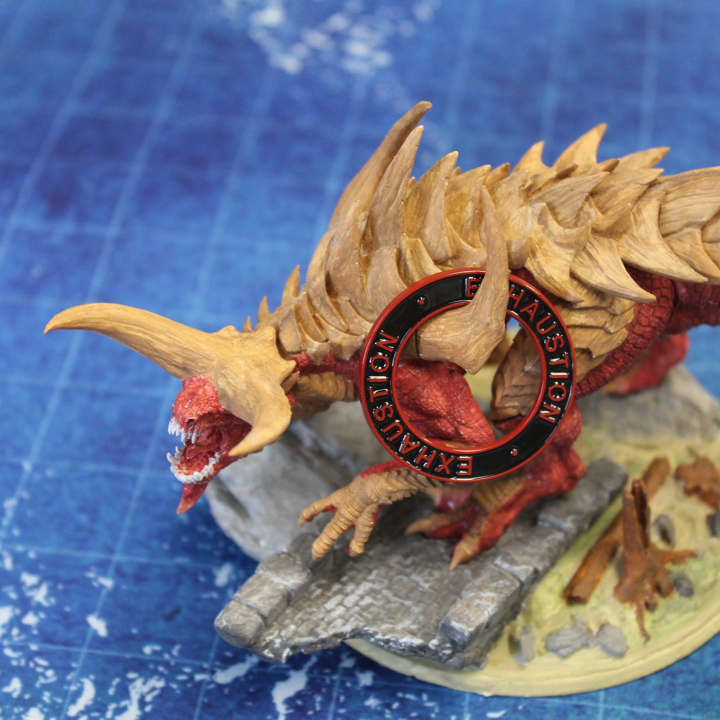 Figure of a tarrasque monster with a black ring with a red rim and red text that says "Exhaustion" on it hanging over one horn on a blue gaming mat.