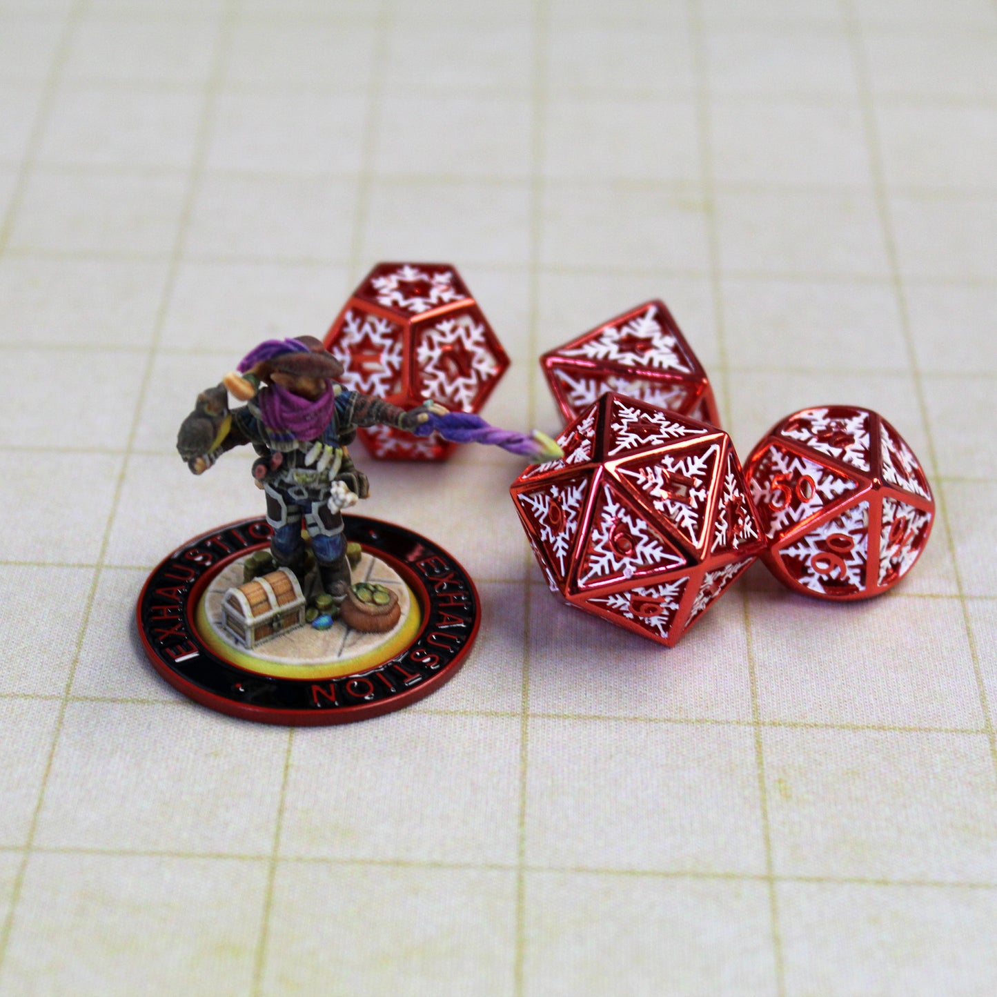 A miniature figure of a spellcasting elf with a black ring with a red rim and red text that says "Exhaustion" on it surrounding the base of the figure sitting next to a few red dice with white snowflake patterns on them.