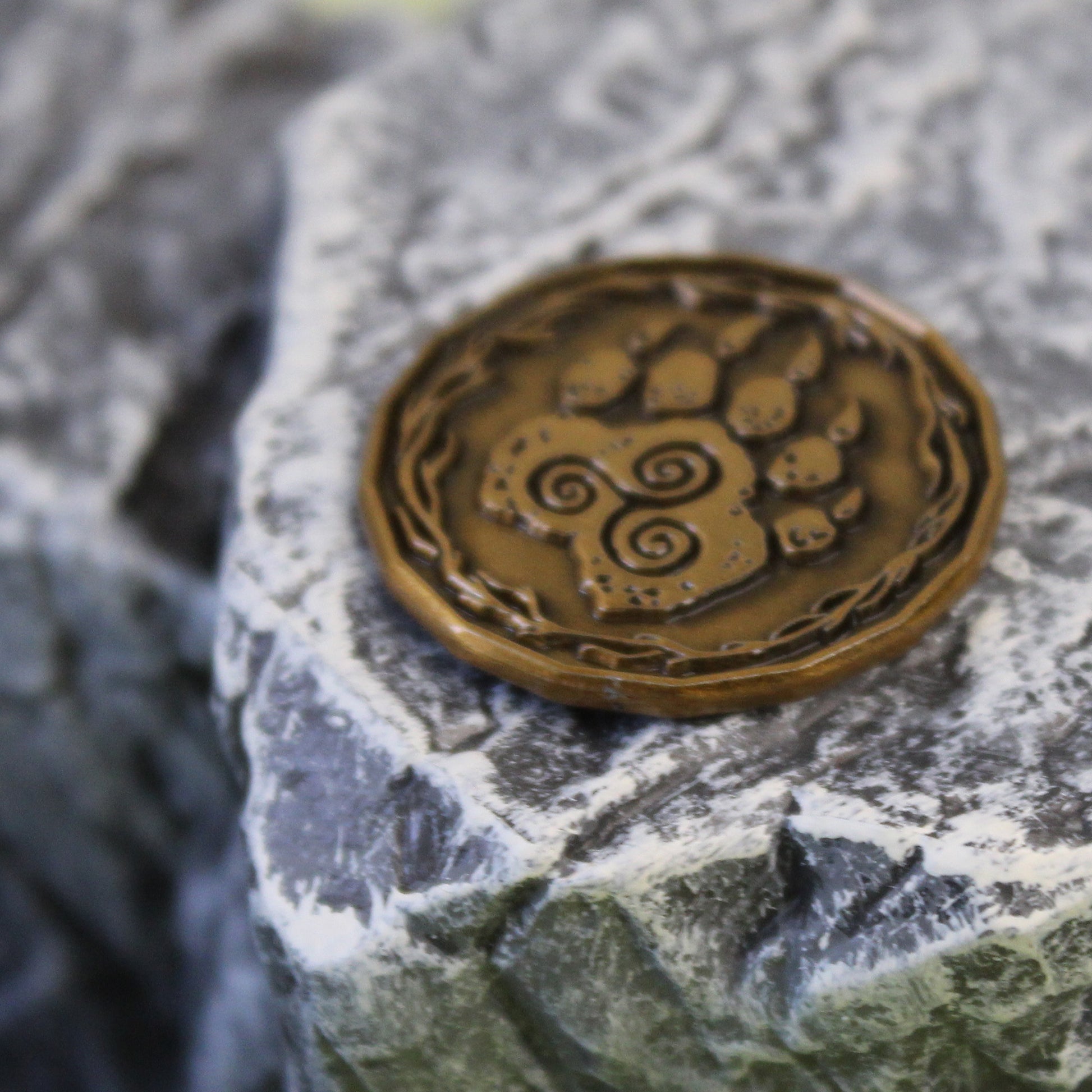 A gold druid character coin on a rocky mountain set piece. The coin is flipped to the "heads" side, depicting a paw with a triple spiral.