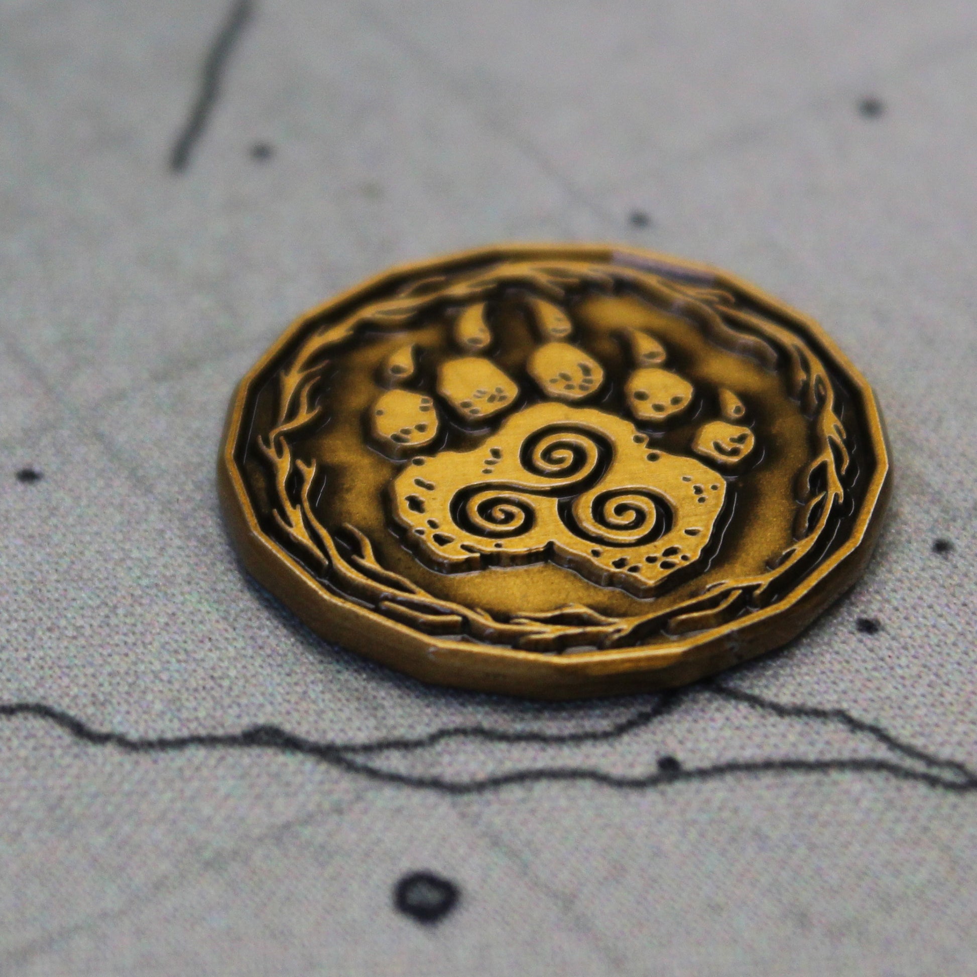 A gold druid character coin on a printed map. The coin is flipped to the "heads" side, depicting a paw with a triple spiral.
