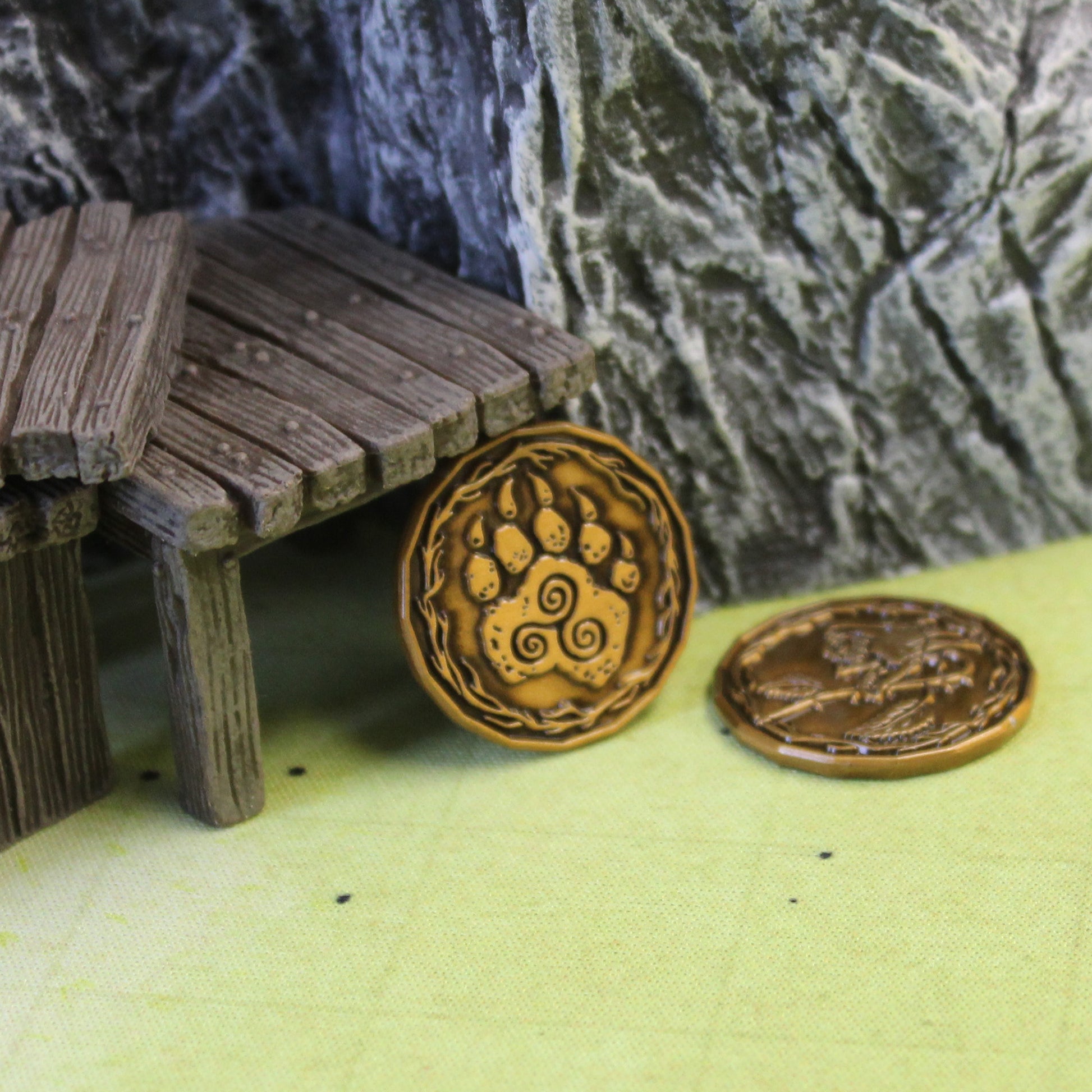 A gold druid character token leaning against a wooden step set piece on a printed map surface. The token is facing the "heads" side, depicting a paw with a triple spiral. Behind the wooden step set piece is a large gray rock set piece. Next to the token is another druid character coin, out of focus, on the "tails" side.