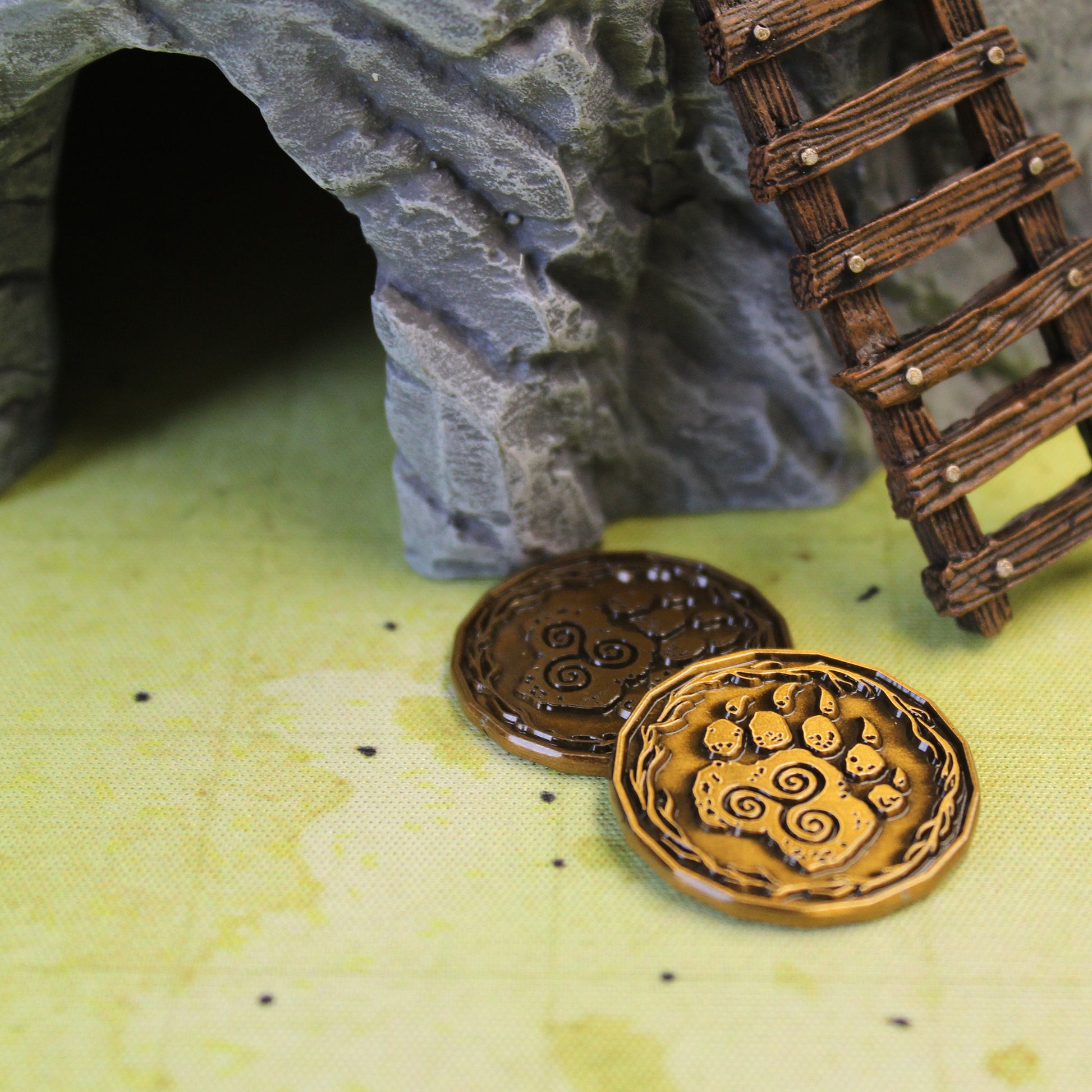Two gold druid character coins on a printed map. One gold character coin is placed slightly on top of the other, and both are flipped to the "heads" side, depicting a paw with a triple spiral. Behind the coins is a rock cave set piece with a wooden ladder set piece leaning on it.