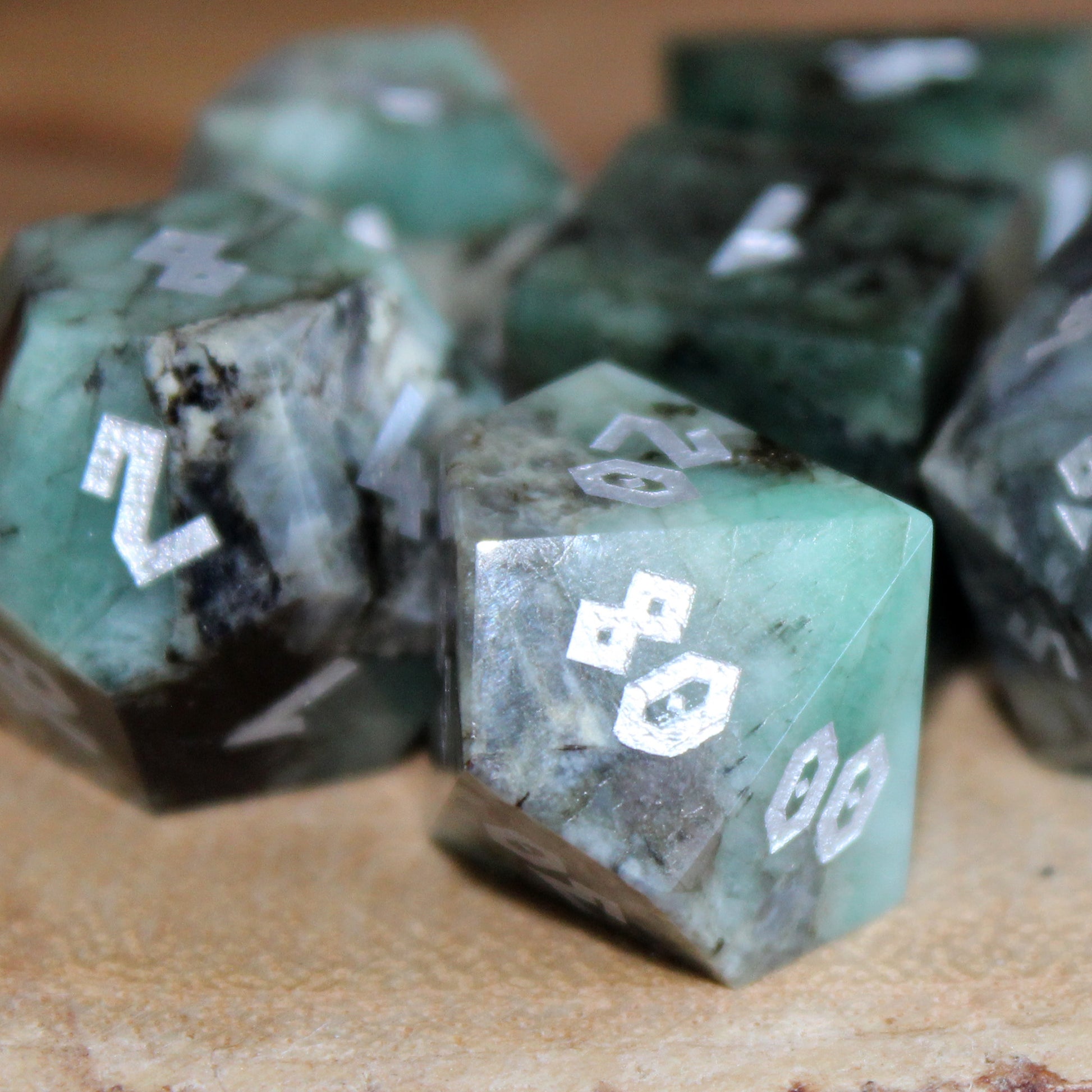 Raw Emerald dice set on a wooden table, with a close-up of the D100, featuring shimmering green stone and silver numbering, perfect for tabletop RPGs.