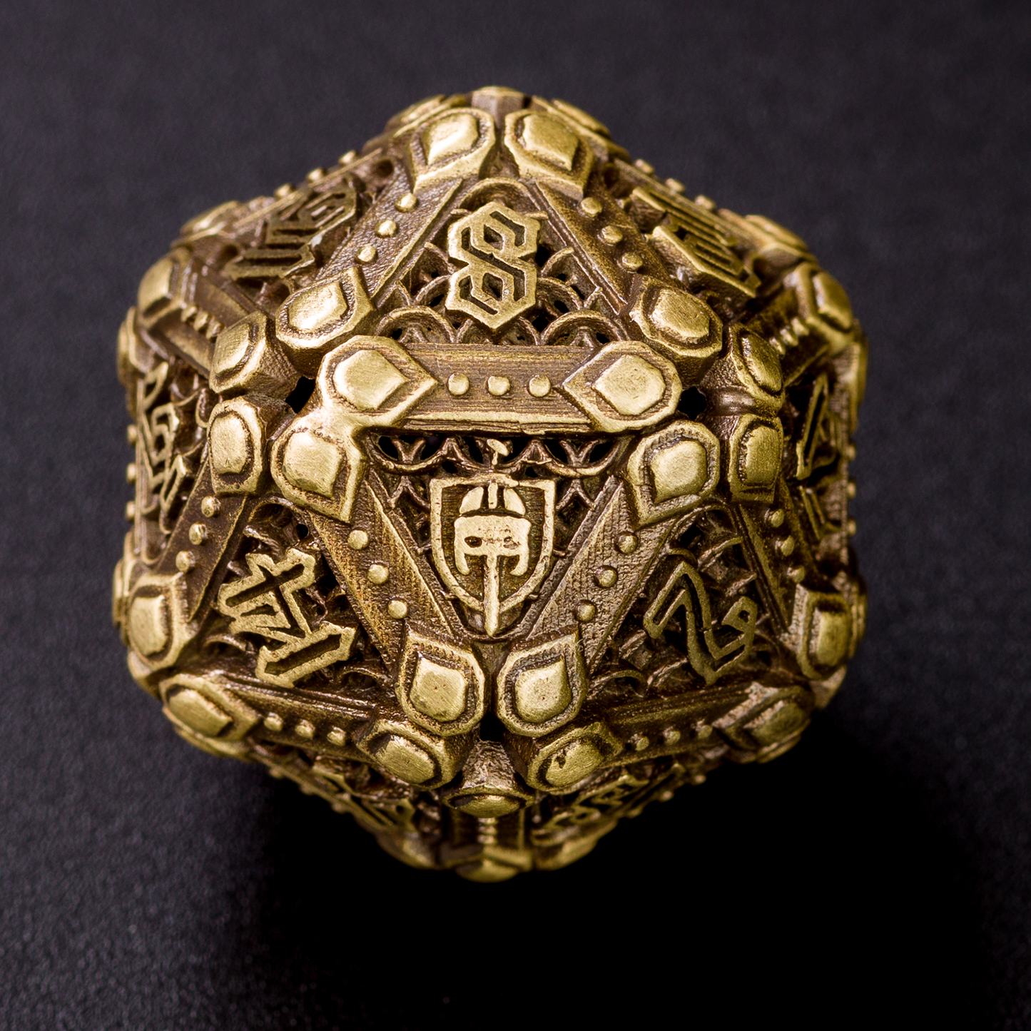 Closeup of bronze fighter-inspired hollow metal twenty-sided die with a chain mail background on each die face, uninked numbers, and a helmet, sword, and shield replacing the high number on the die on a back background