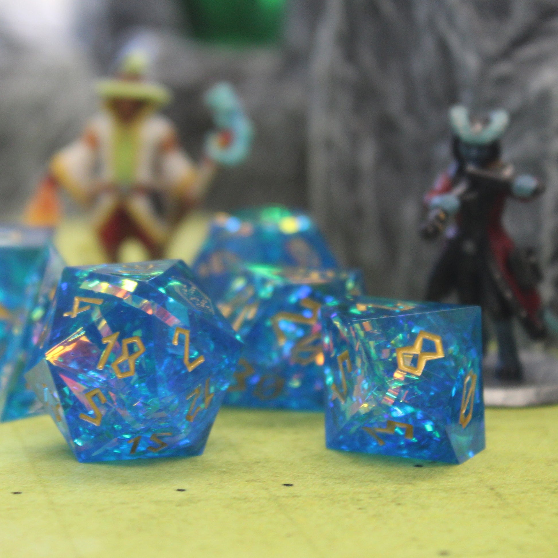 StarGlow Blue dice set arranged in a pile on a green game mat, with only the D20 and a D10 in focus in the front. Behind the dice are two mini figures and a gray rocky wall set piece.
