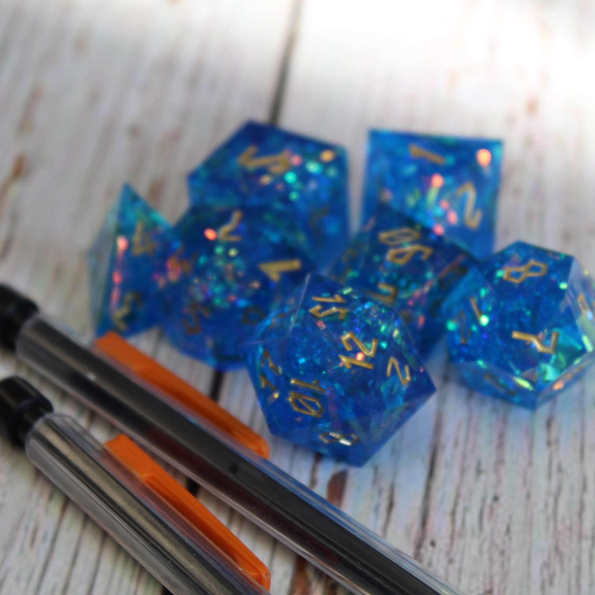 StarGlow blue dice arranged in a pile on a white wood surface. In front of the dice are two mechanical pencils, only the back half of the pencils can be seen within the image.