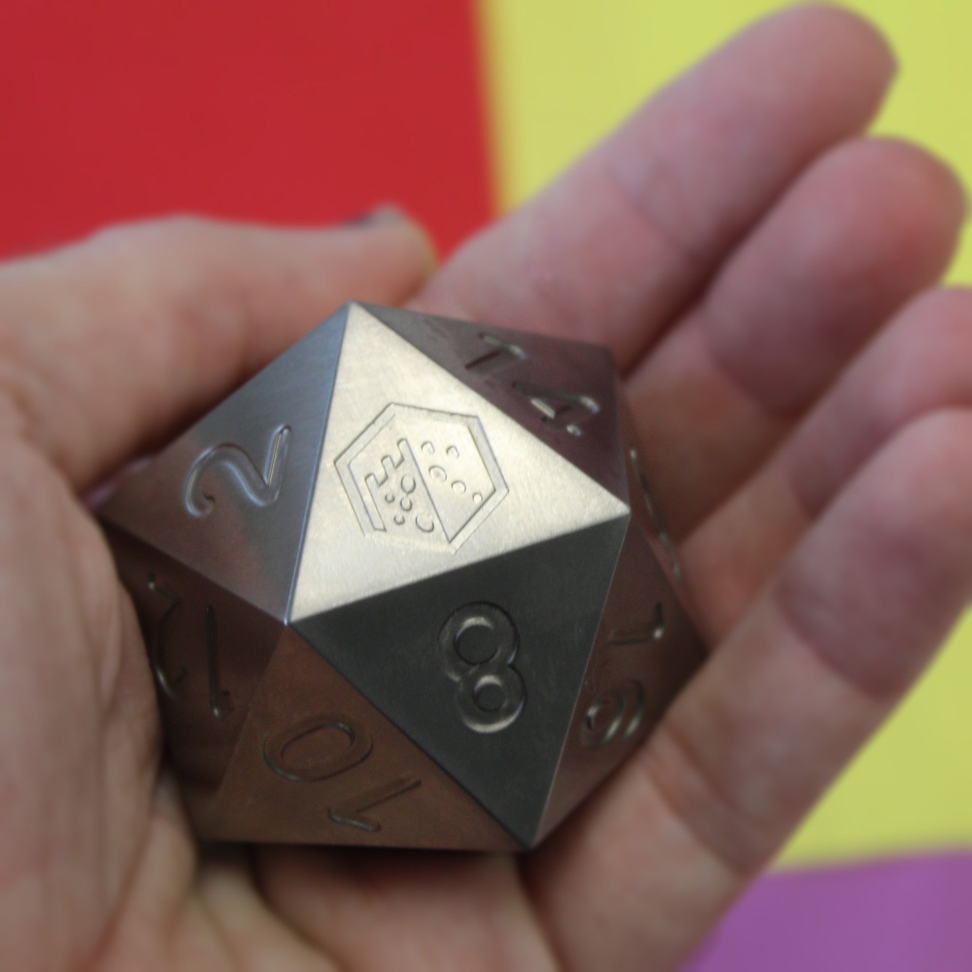 An oversized silver-colored metal twenty-sided die with a Dice Dungeons logo in place of the number twenty in a person's palm.