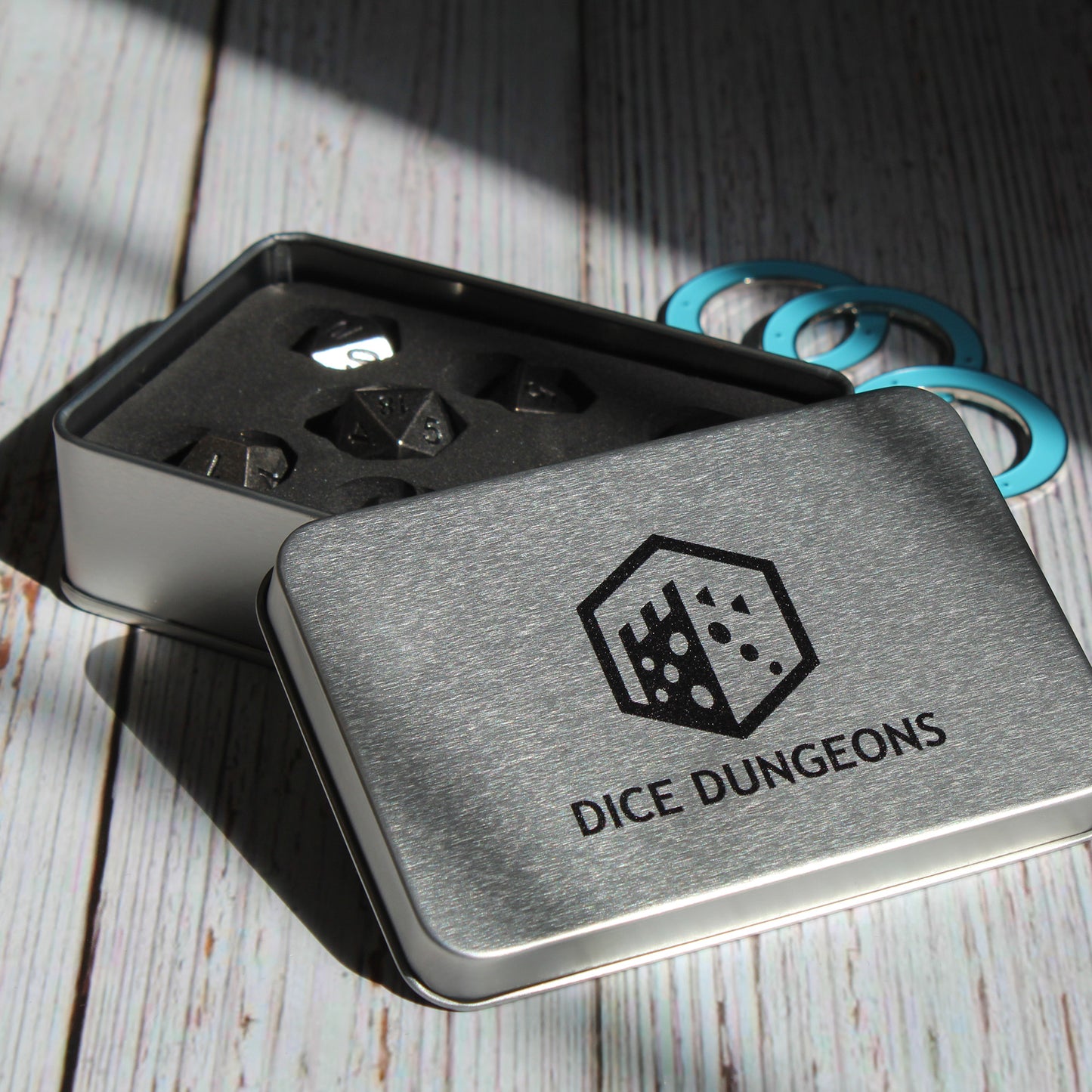 Primordial silver dice set inside of a Dice Dungeons silver display box on a light wooden surface. The lid of the box is leaning against the base so that the dice can be seen positioned inside and next to the box is three teal condition rings.