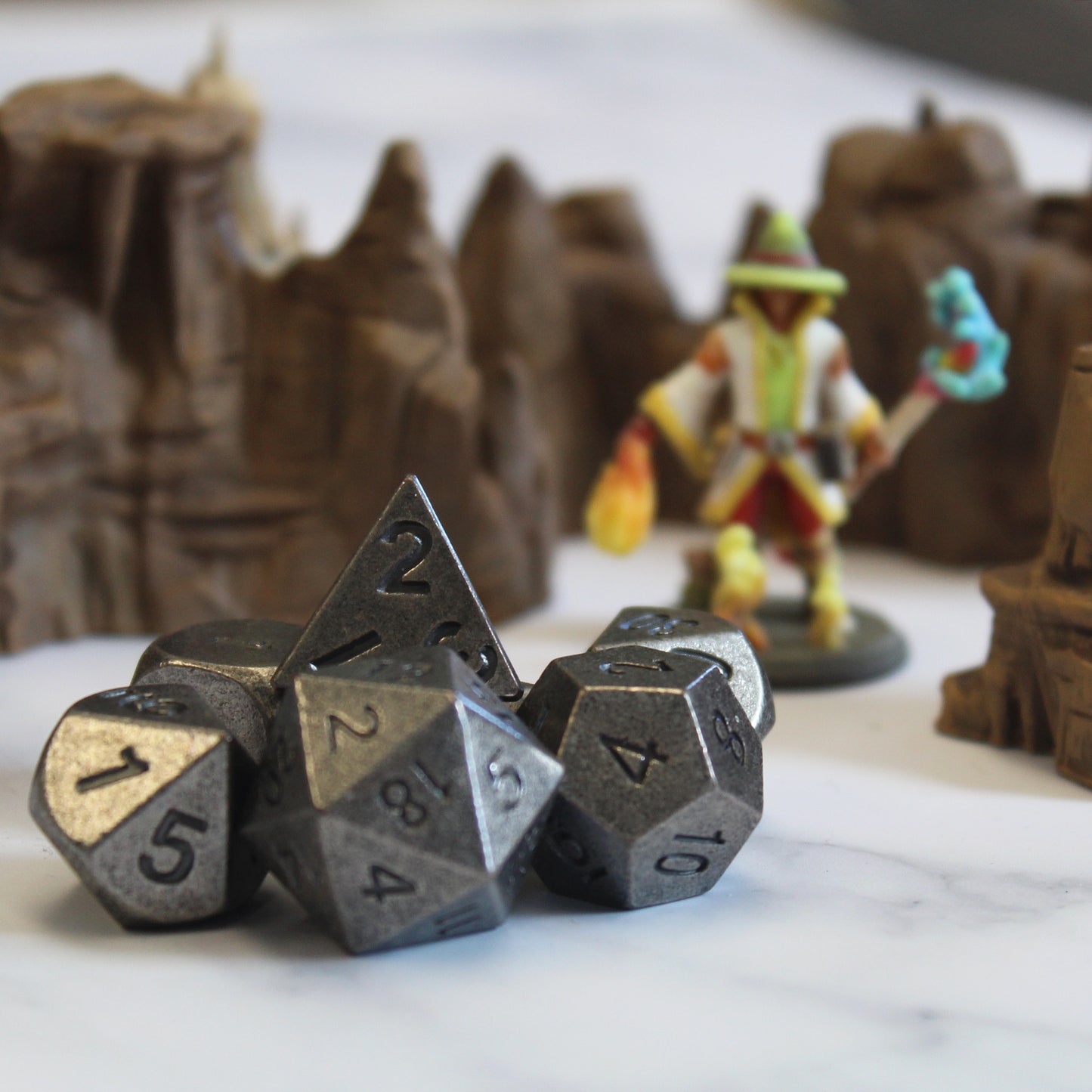 Primordial silver dice set arranged on a white surface, with rocky desert set pieces and a mini figure out of focus behind them