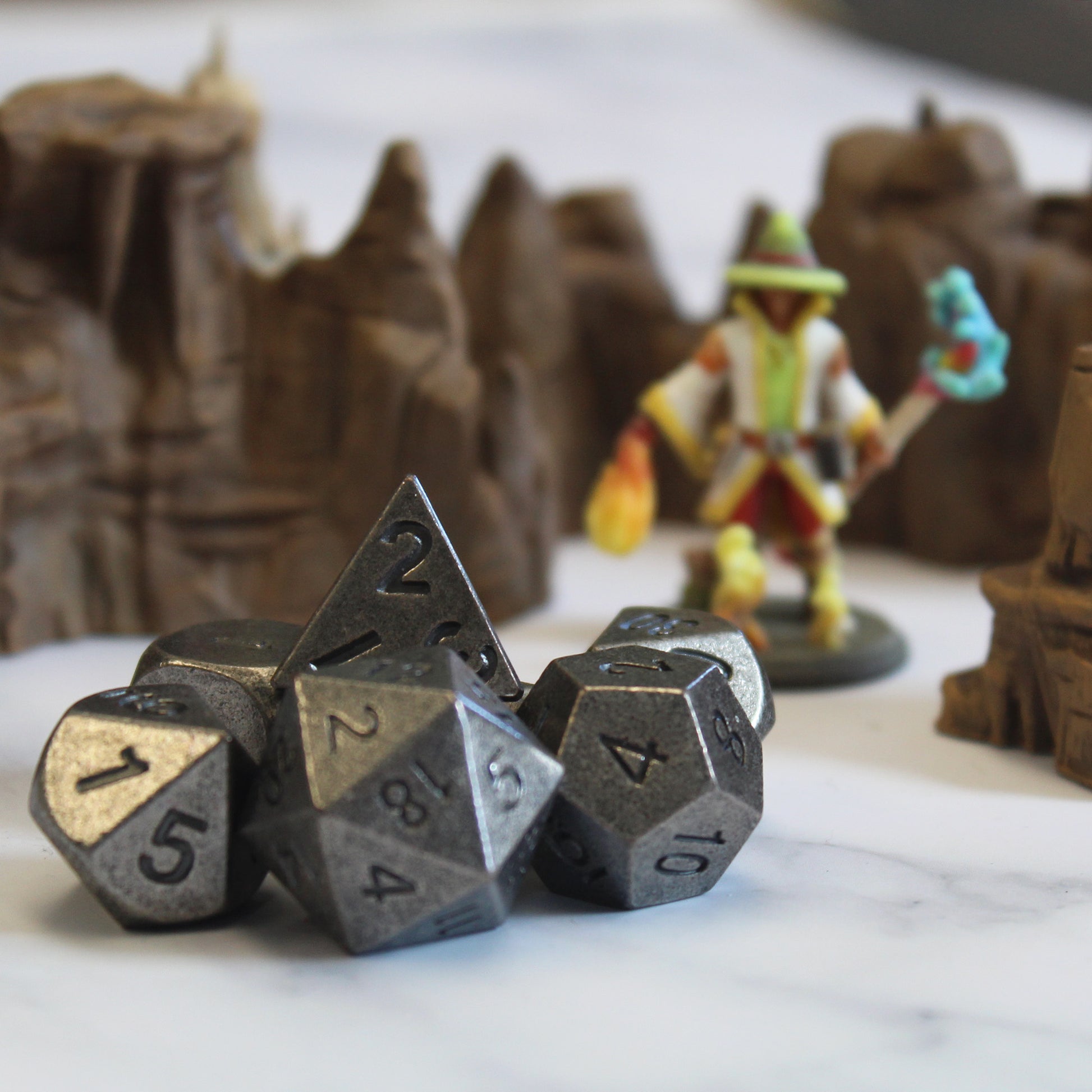 Primordial silver dice set arranged on a white surface, with rocky desert set pieces and a mini figure out of focus behind them