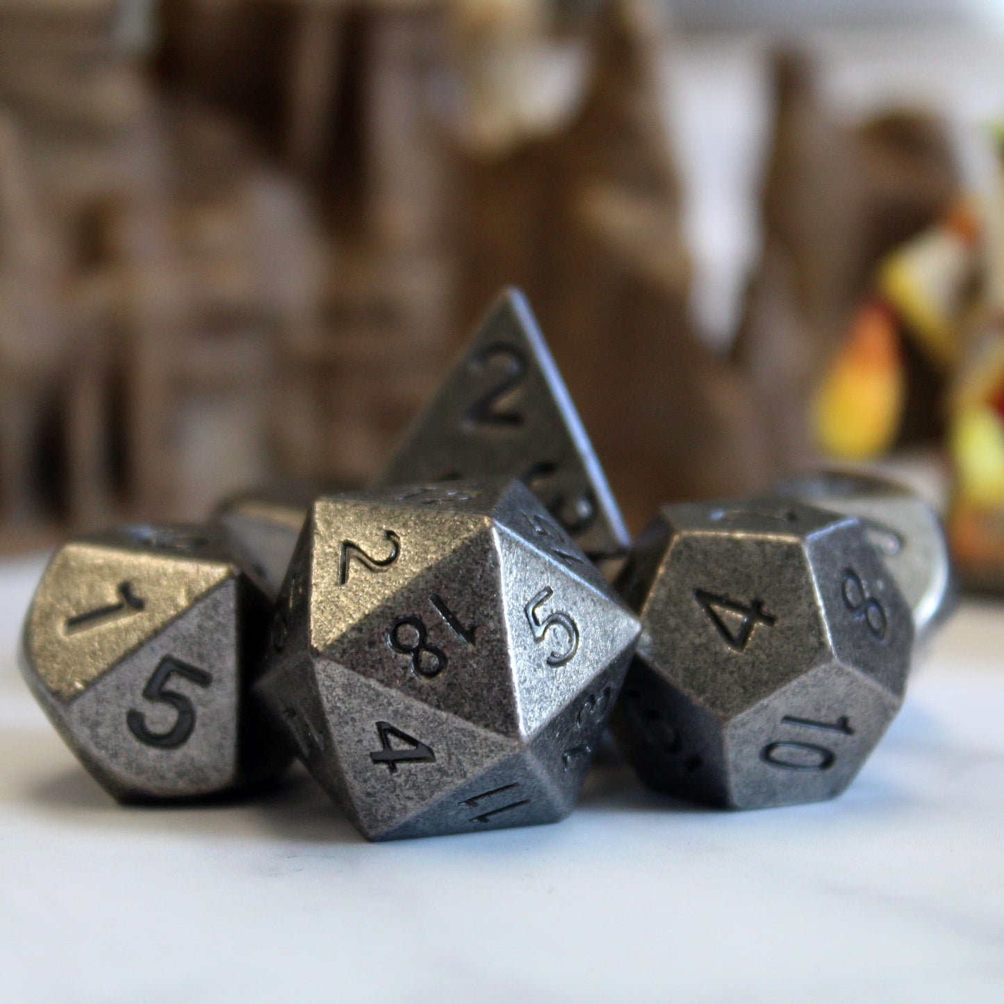 Close-up of Primordial silver dice set arranged on a white surface, with tan set pieces behind them. The close-up showcases the antique silver faces and black numbering.