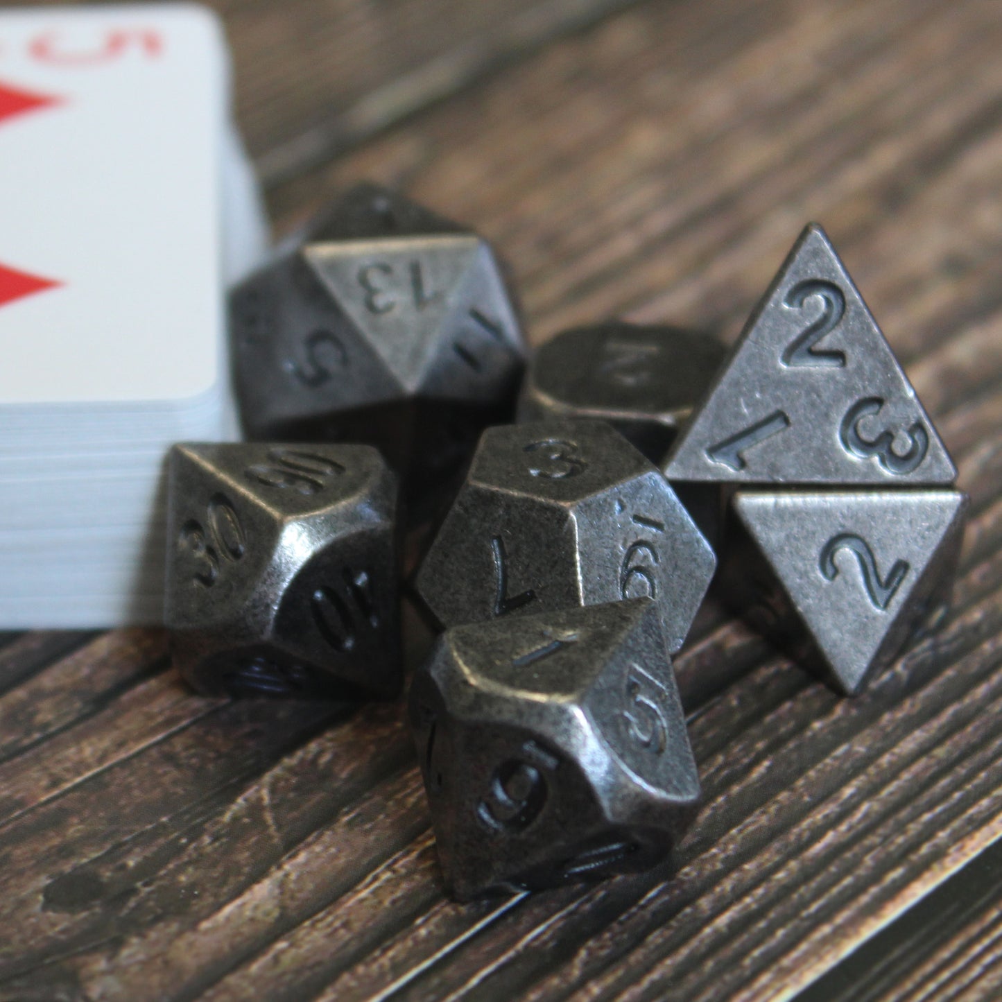 Primordial silver dice set arranged next to a stack of playing cards with the red five of diamonds on top, on a dark wooden surface