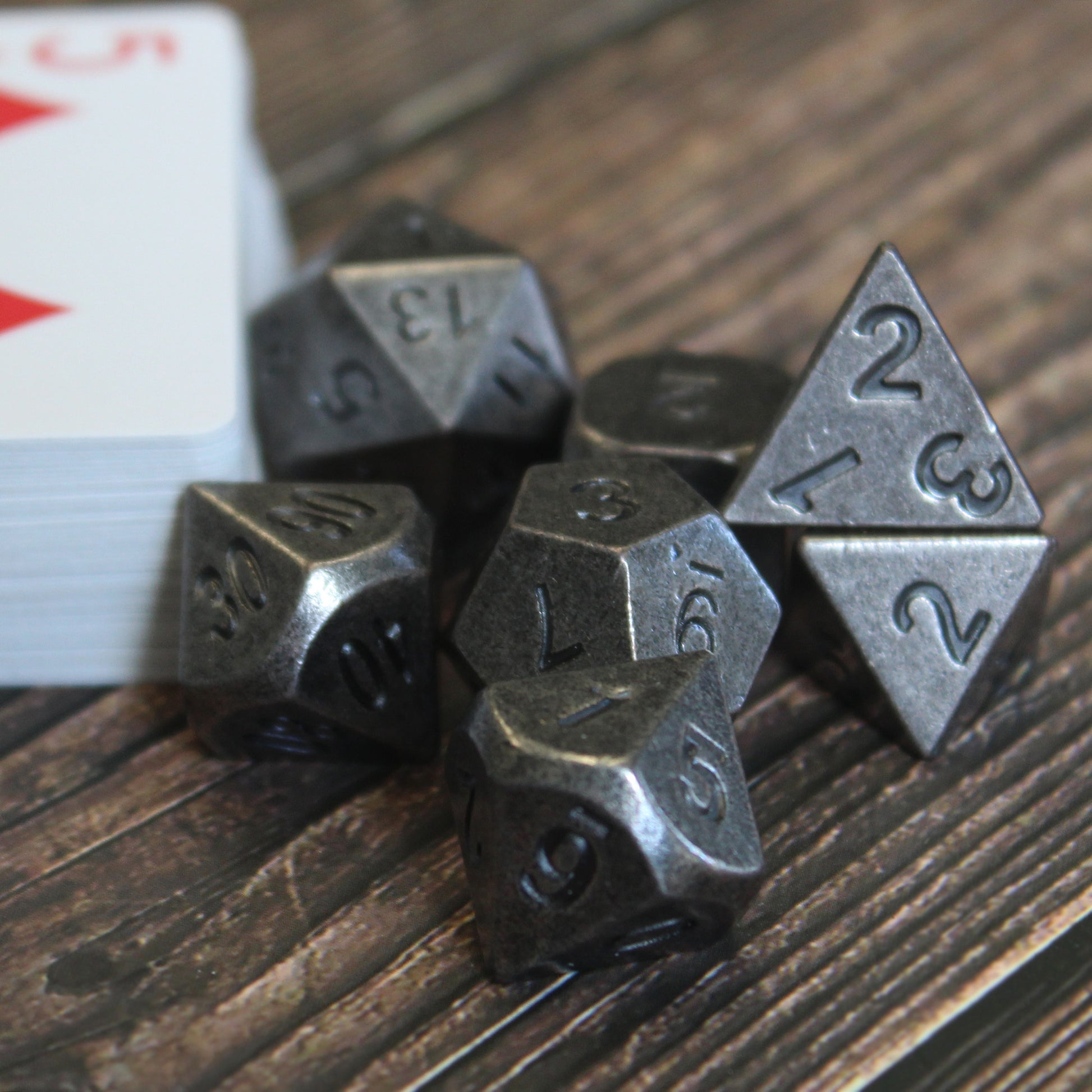 Primordial silver dice set arranged next to a stack of playing cards with the red five of diamonds on top, on a dark wooden surface