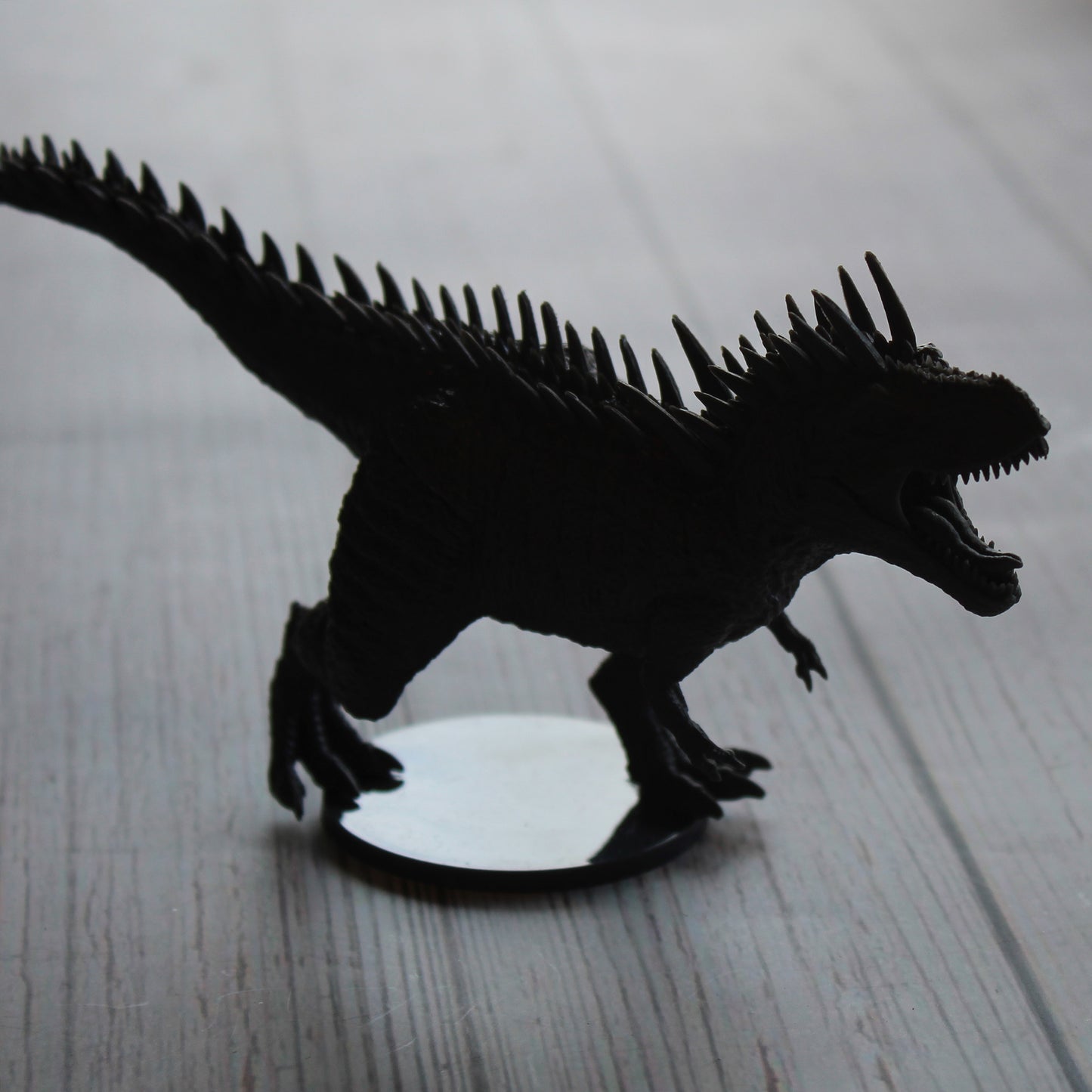 grey miniature figure t-rex in shadow, mouth open and facing to the right of the image. On a white wood paneled surface.