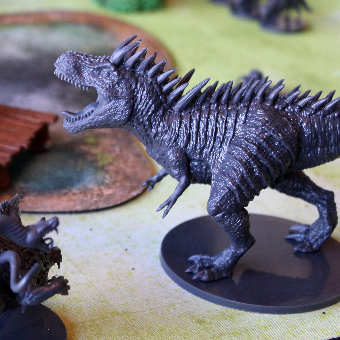 grey miniature figure t-rex shown in game play, surrounded by other figurines and items. T-rex figurine is standing, mouth open, facing to the left of the image.