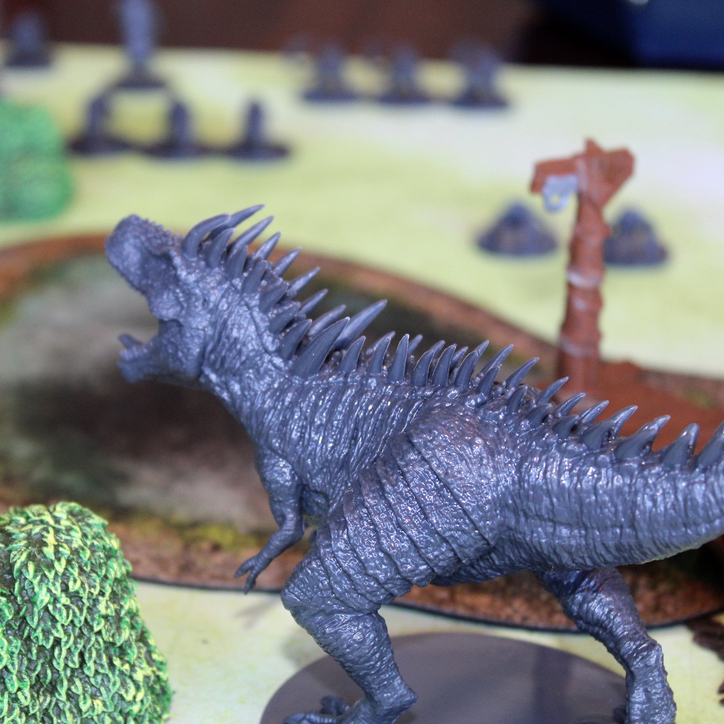 grey miniature figure t-rex shown in game play, surrounded by other figurines and items. T-rex figurine is standing, mouth open, facing to the left of the image.