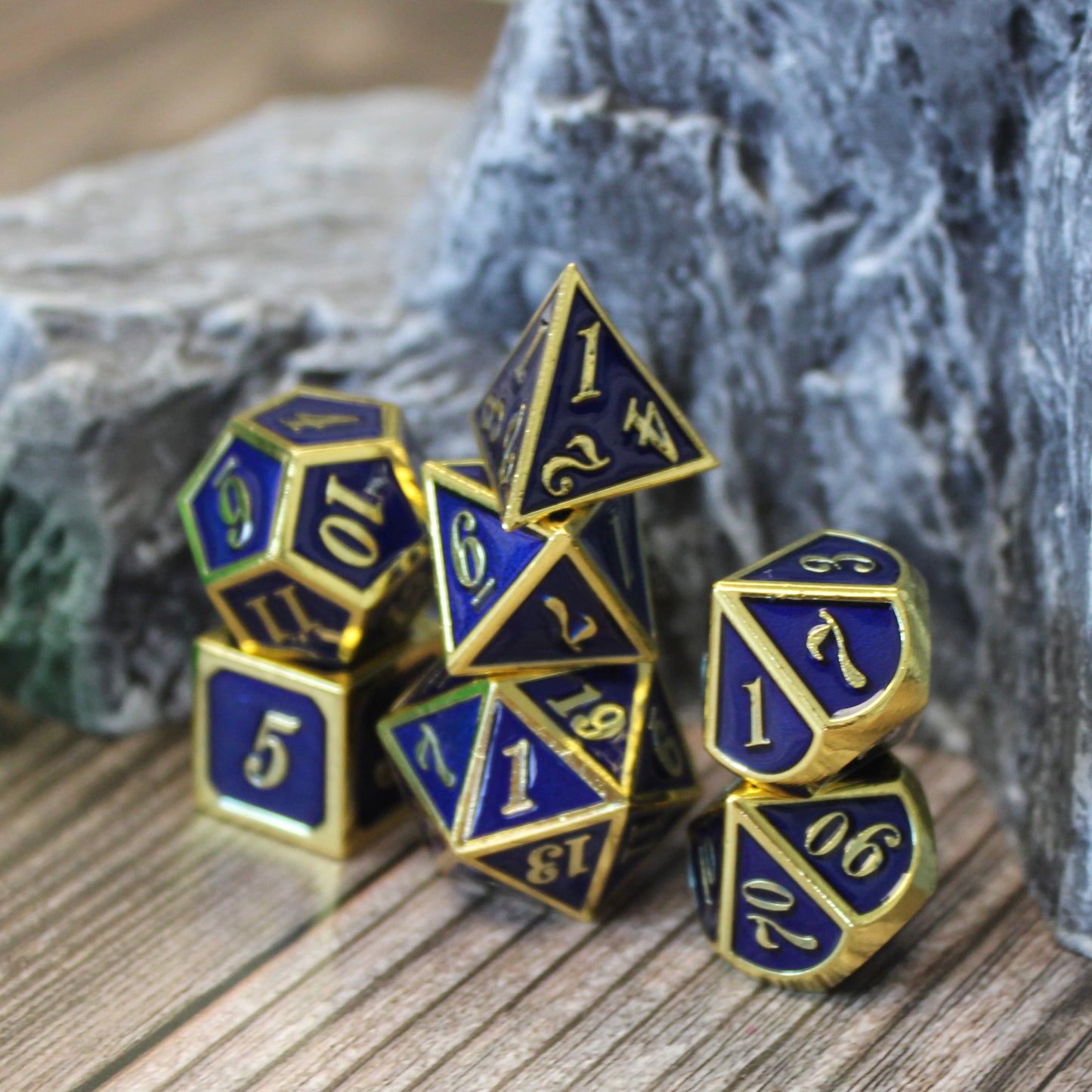 Imperial blue and gold dice set displayed in three stacks on top of a wooden surface, with a rocky set piece behind them.