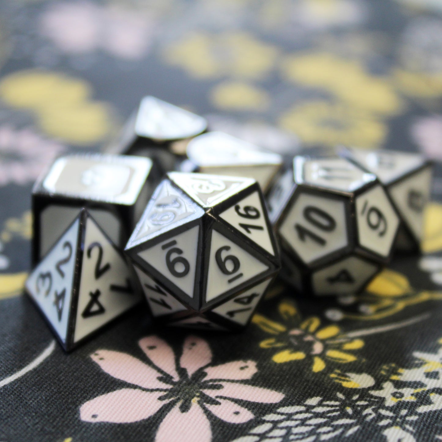 Metal Imperial White dice set scattered on a black fabric with yellow and pink flowers. The dice have a white face and black numbers and framing.