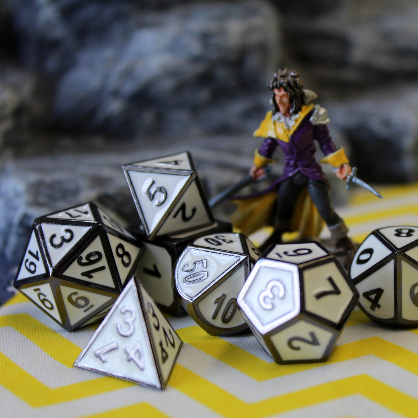 Imperial White dice set scattered on a yellow and white chevron patterned surface, with a mini figure and a rocky set piece positioned behind them
