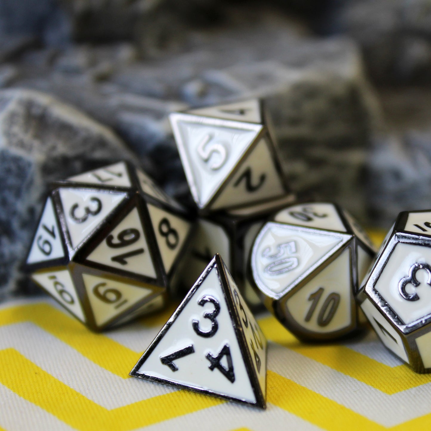 Metal Imperial White dice set scattered on a yellow and white chevron patterned fabric, with a rocky set piece behind them