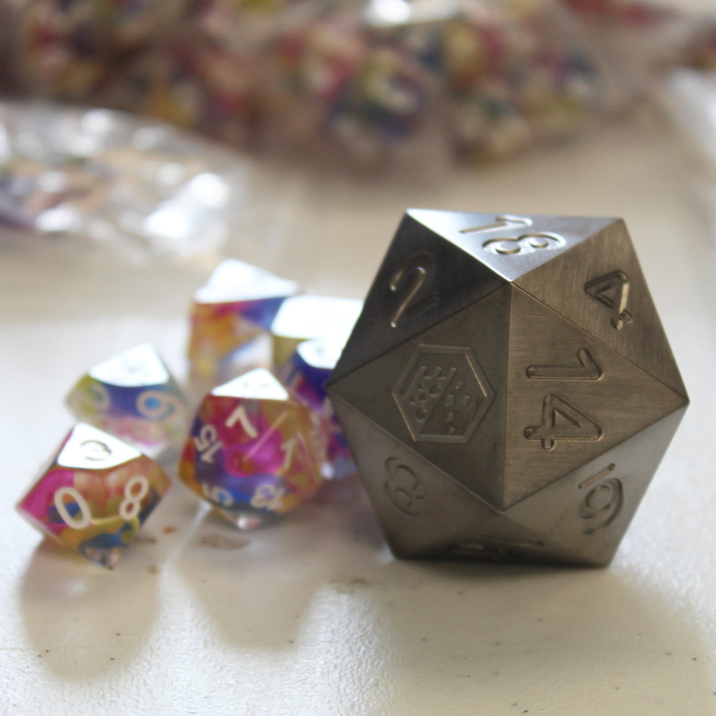 An oversized silver-colored metal twenty-sided die with a Dice Dungeons logo in place of the number twenty sitting next to a set of normal-sized rainbow colored dice on a white surface.