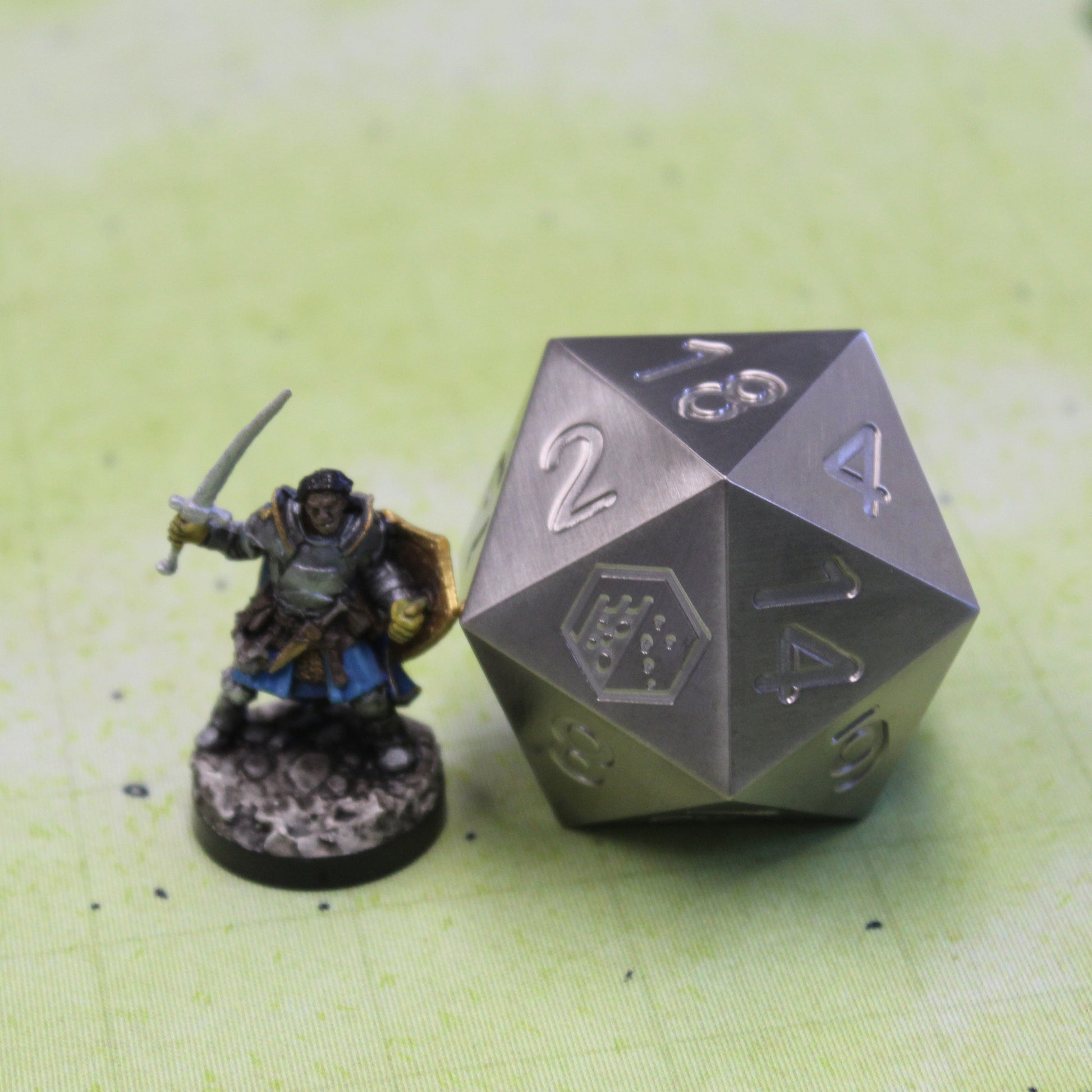 An oversized silver-colored metal twenty-sided die with a Dice Dungeons logo in place of the number twenty sitting next to a miniature figure on a green gridded gaming mat.
