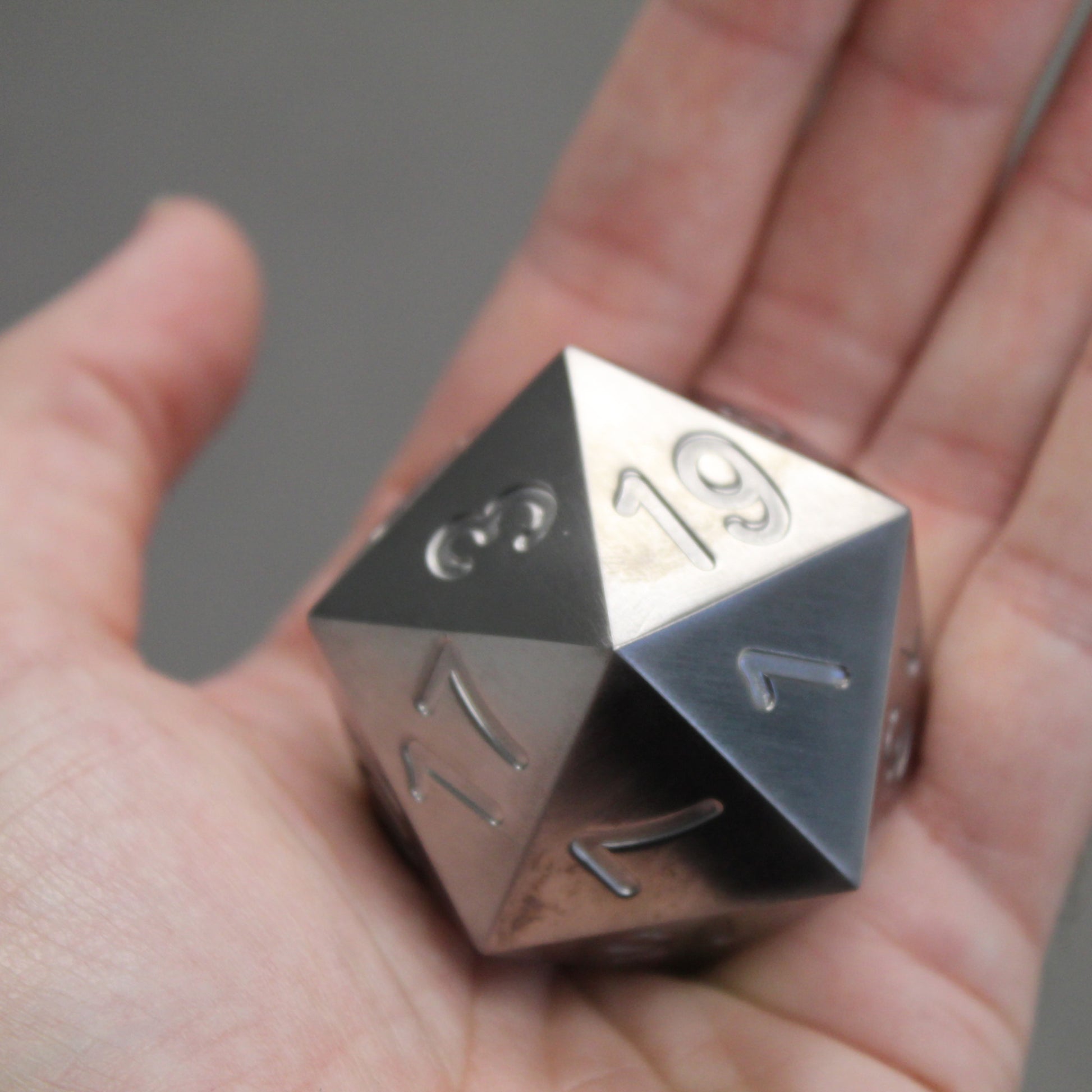 An oversized silver-colored metal twenty-sided die with a Dice Dungeons logo in place of the number twenty in a person's palm