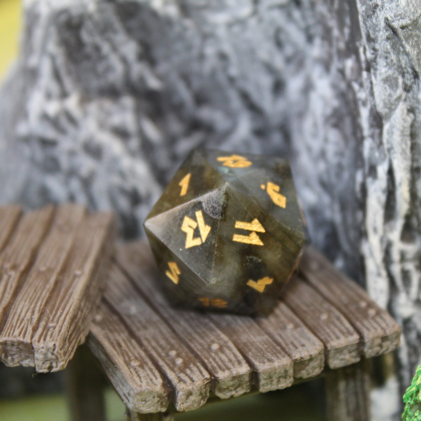 D20 Labradorite Die on a wooden step set piece in front of a gray rocky background set piece.