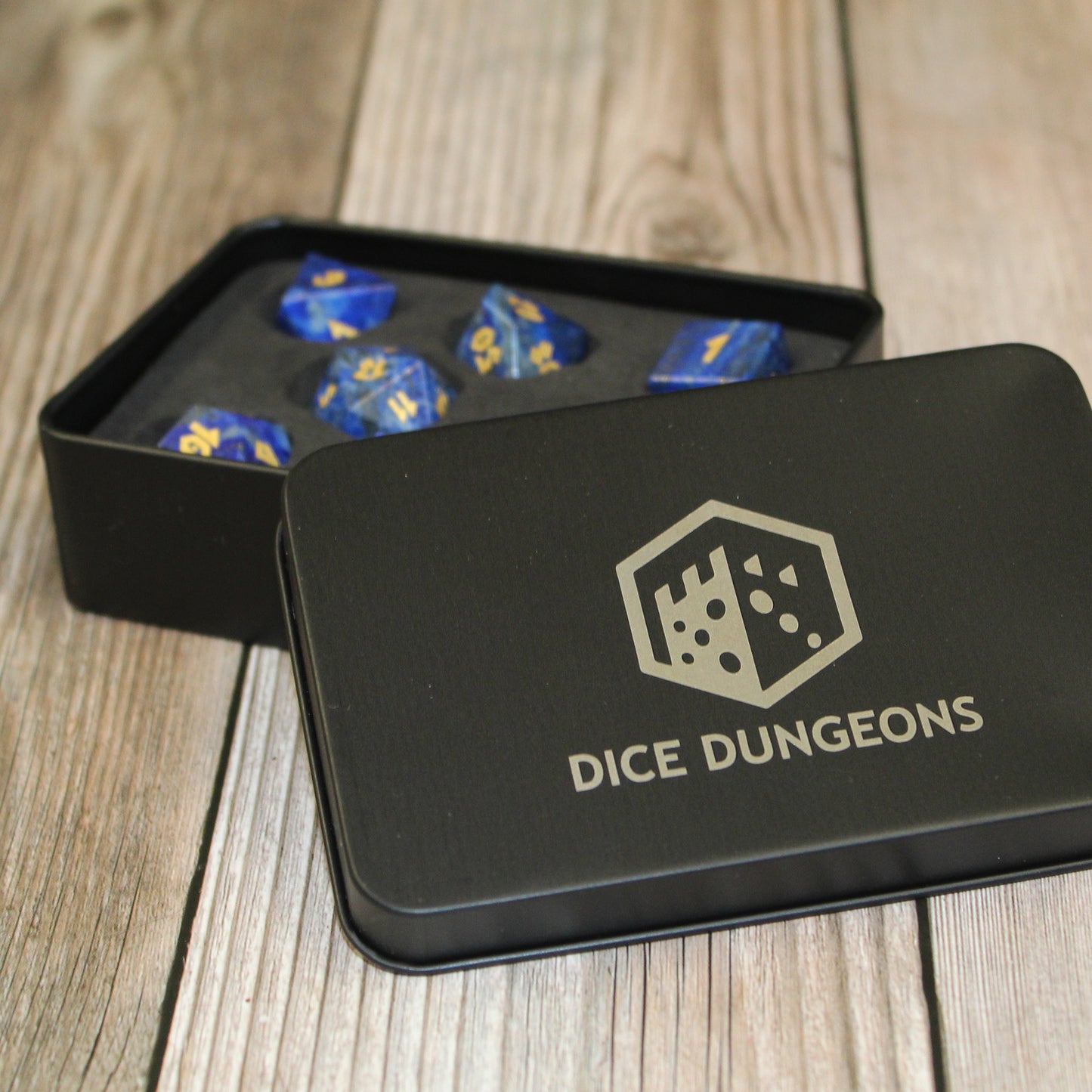 Lapis Lazuli dice set inside of a Dice Dungeons black display box on a light wooden surface. The box is open so that the dice can be seen positioned inside the box, with the lid leaning against the base.